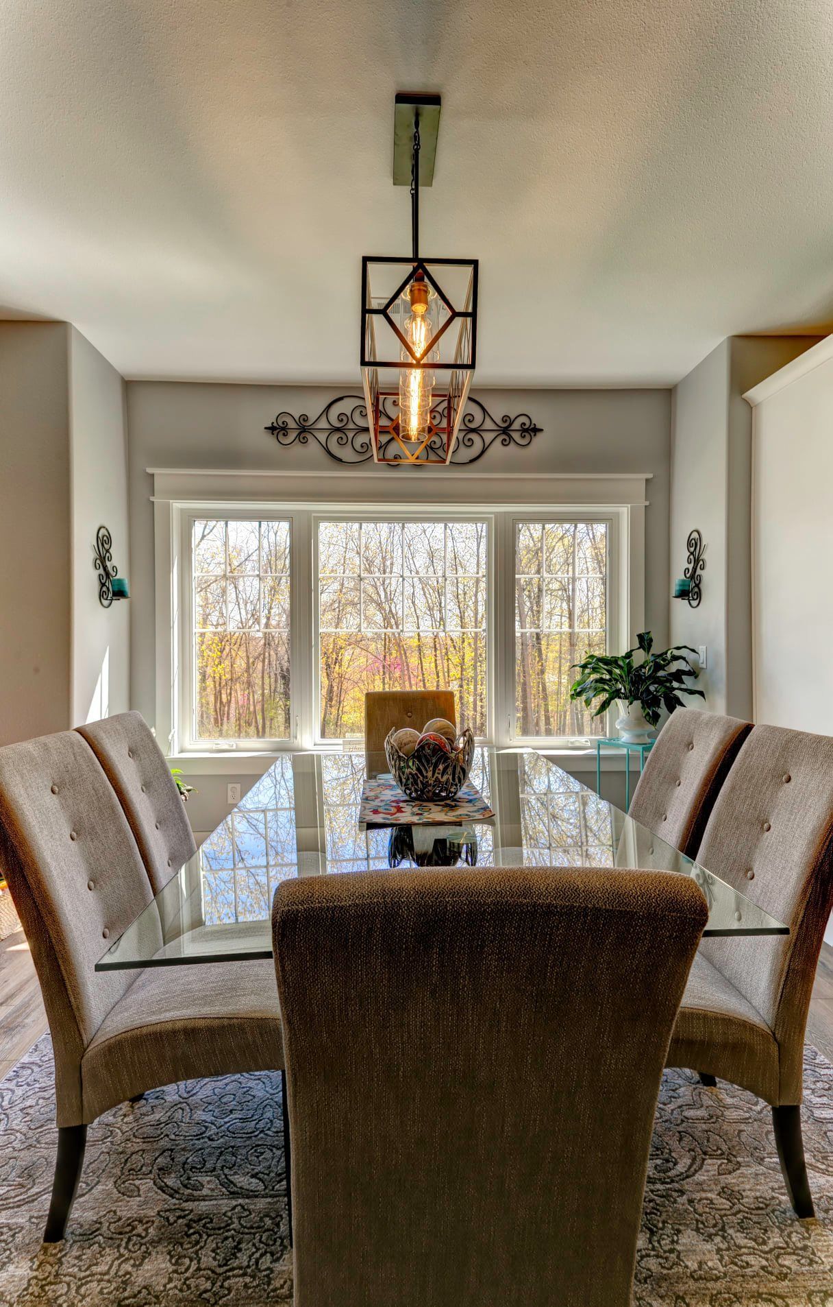 Modern dining room with upholstered chairs, glass table, chandelier, and large windows overlooking trees