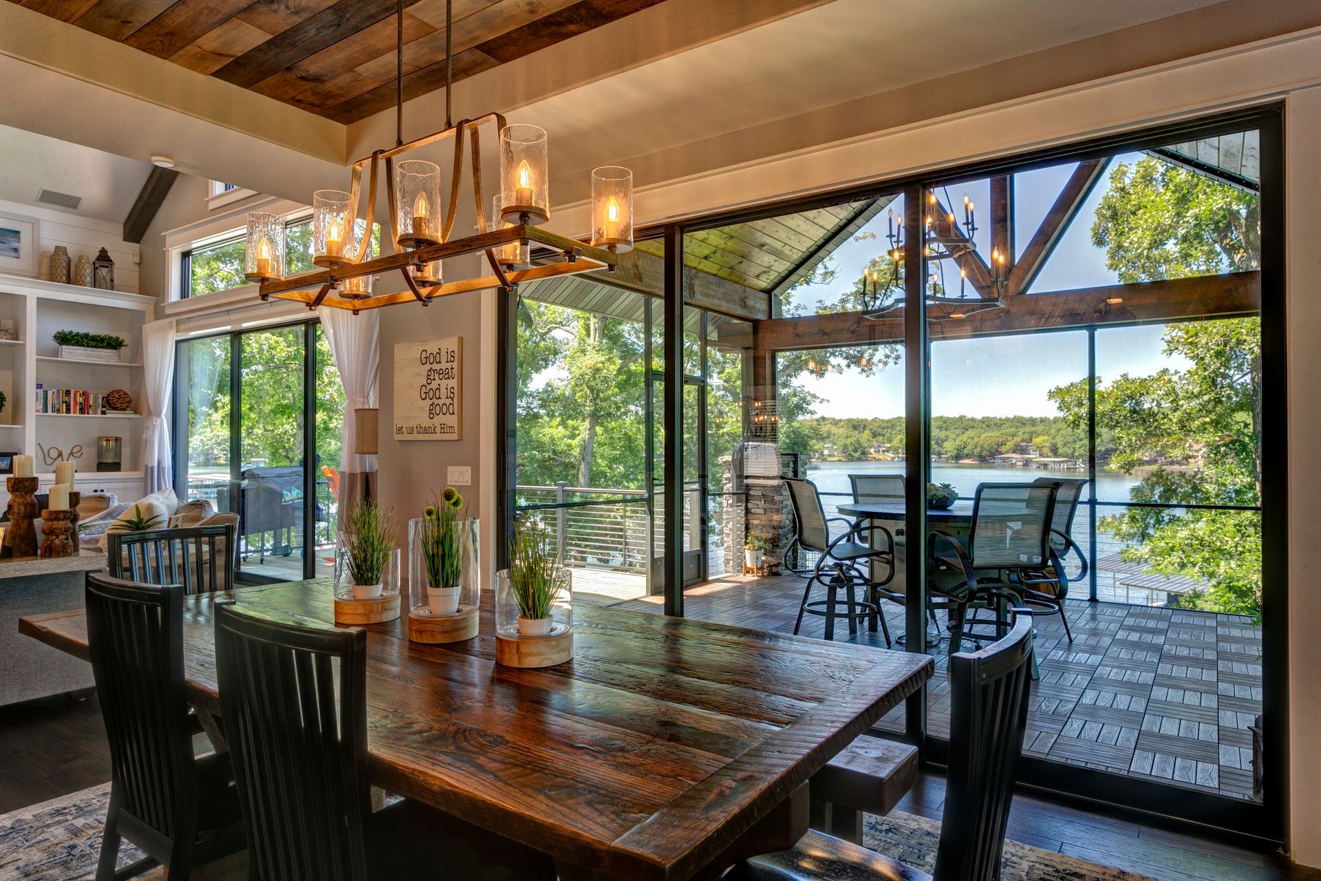 Open-air dining room with rustic wood table, chandelier, and glass doors overlooking a lake and patio.