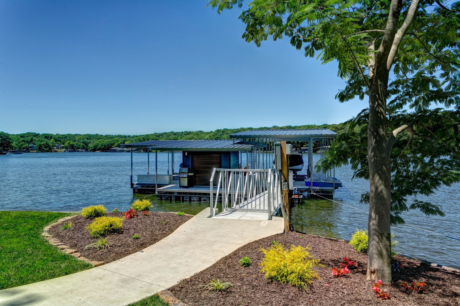 Wooden dock on a lake with a blue canopy, paved path, and landscaped flower beds under clear sky