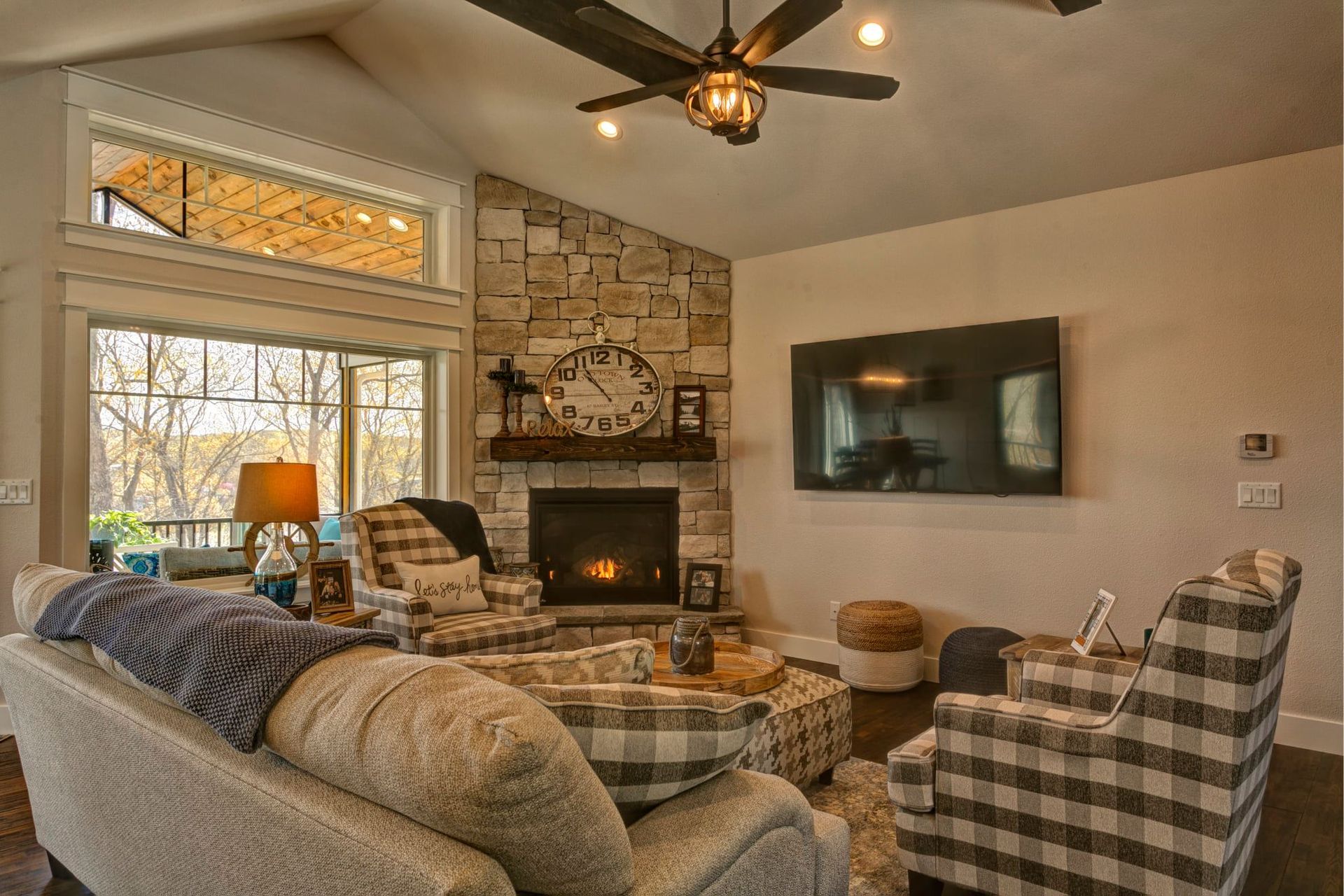 Cozy living room with stone fireplace, plaid chairs, wall-mounted TV, and large windows