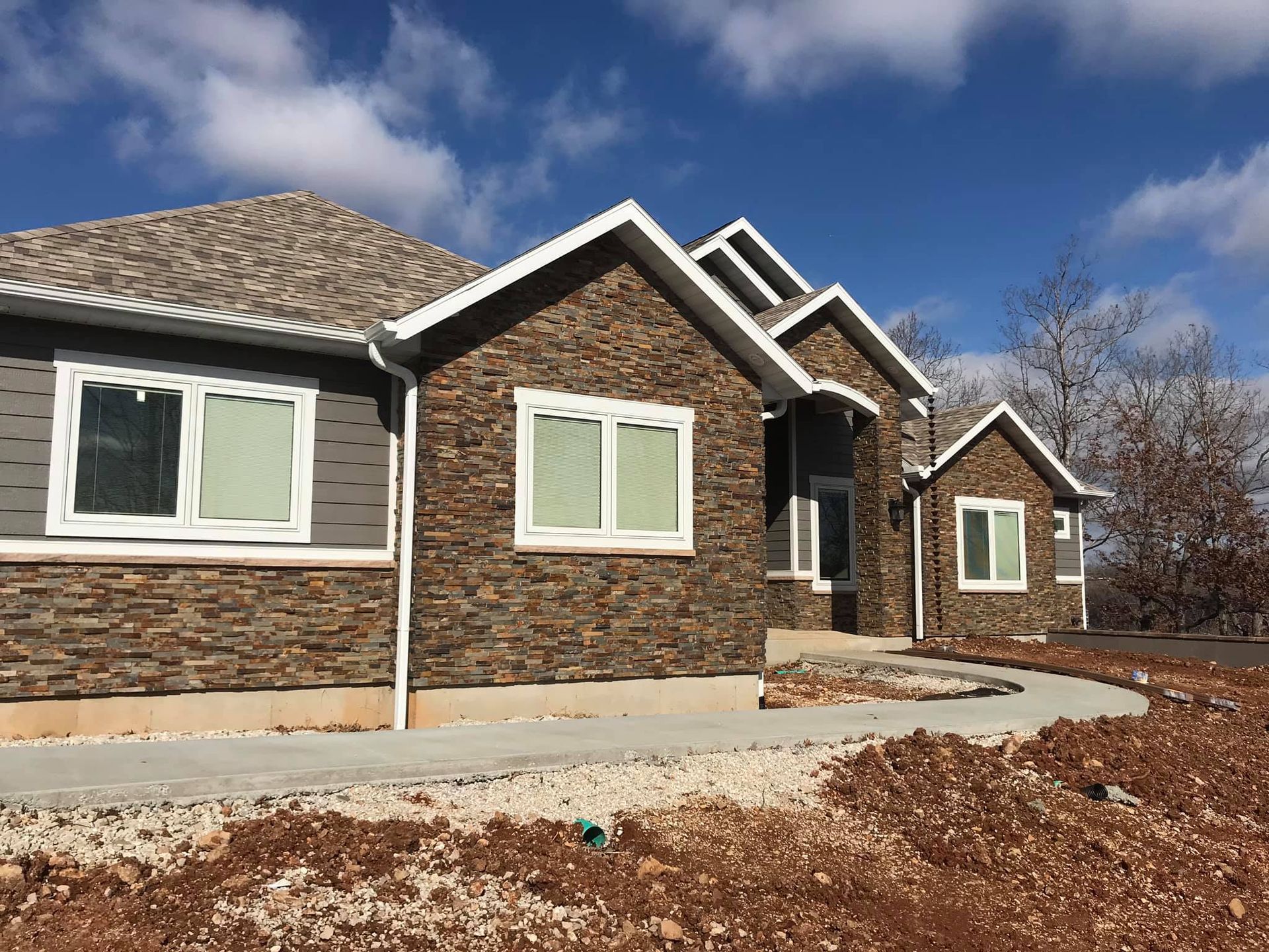 Single-story house with stone and gray siding, front walkway, and leaf-covered yard under a blue sky
