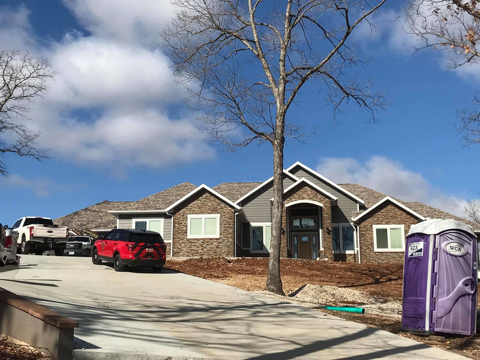 Suburban house under construction with a red truck, bare trees, and portable toilets in front