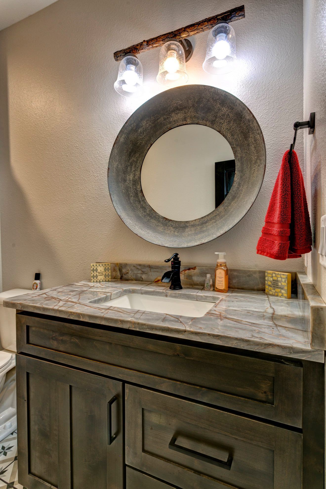 Bathroom vanity with round mirror, three lights, marble countertop, and red hand towel