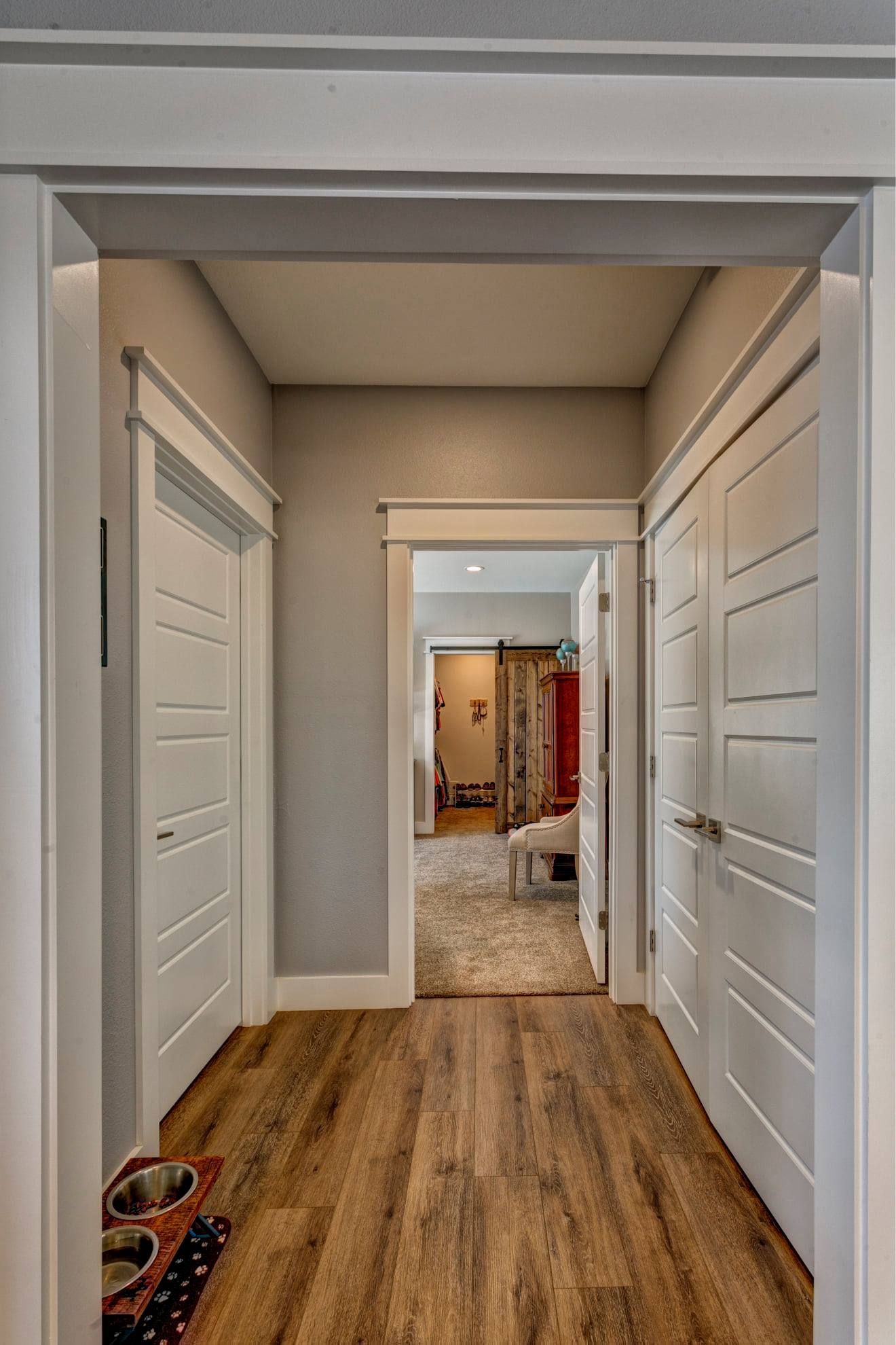 Narrow hallway with white doors, wood floor, and a view into another room beyond.
