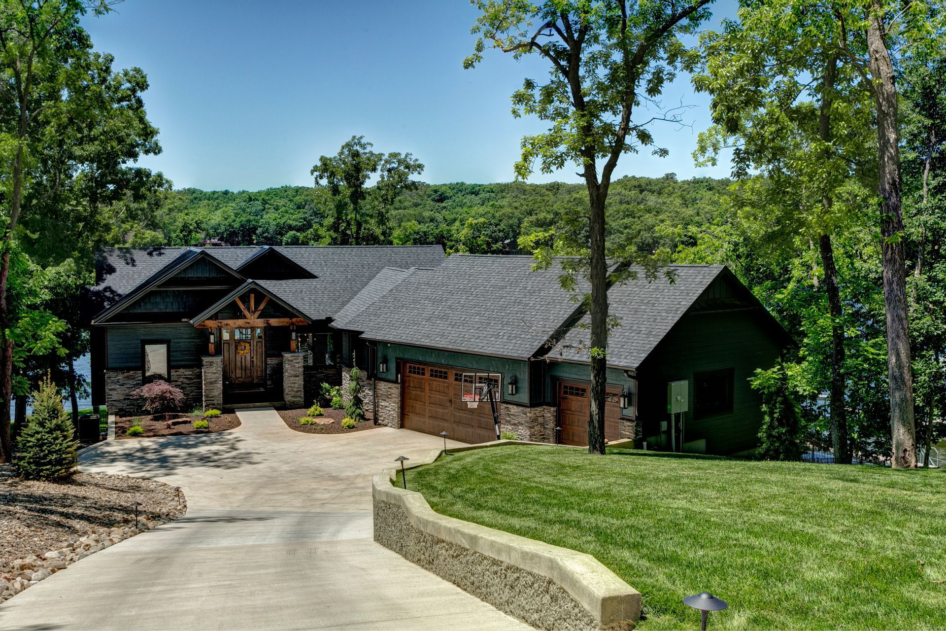 Large modern house with stone driveway, green lawn, and trees on a sunny day