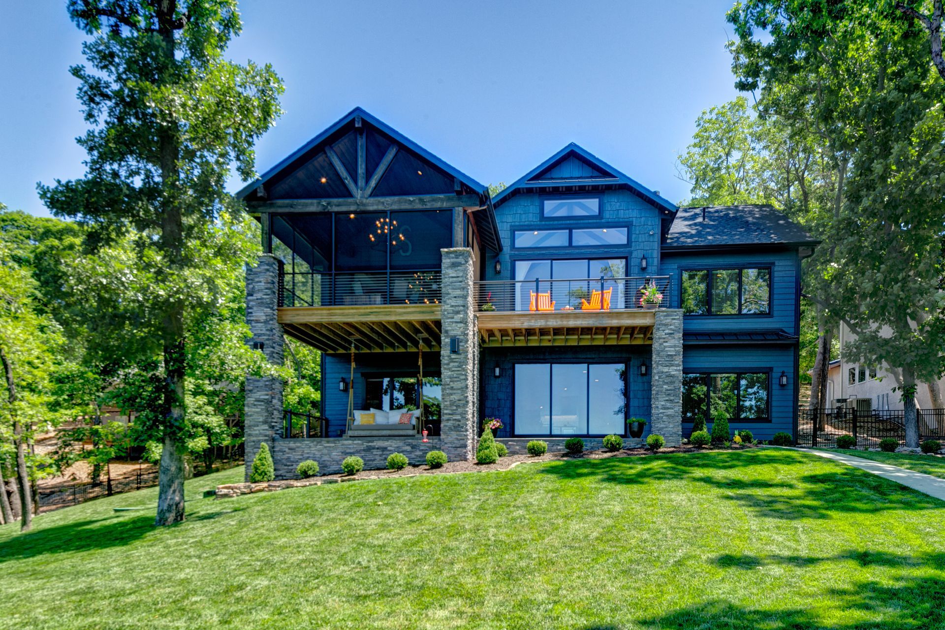 Large modern black house with balconies on a green lawn, surrounded by tall trees under a blue sky