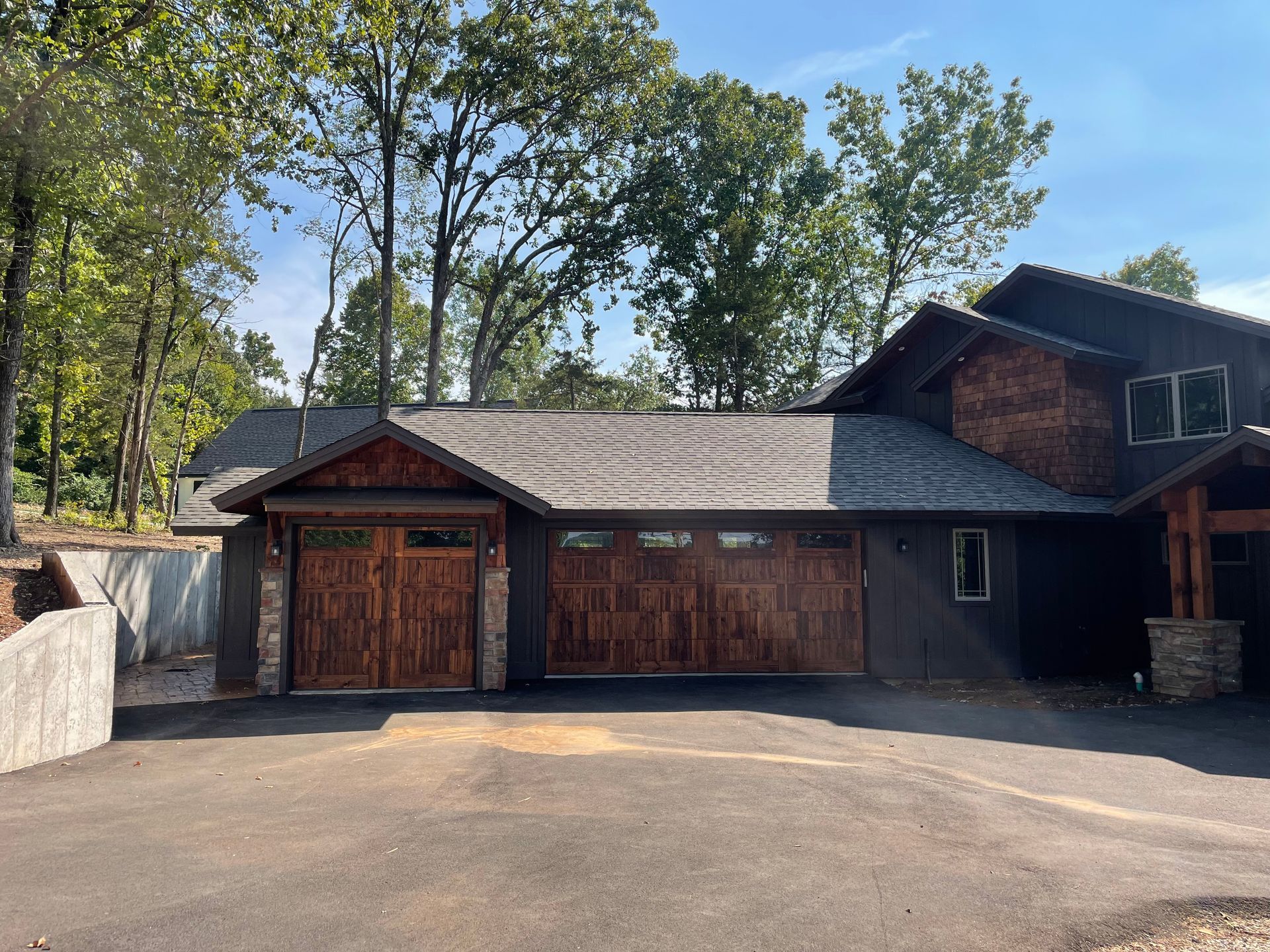 Wooden house with two garage doors, stone accents, and a paved driveway surrounded by trees