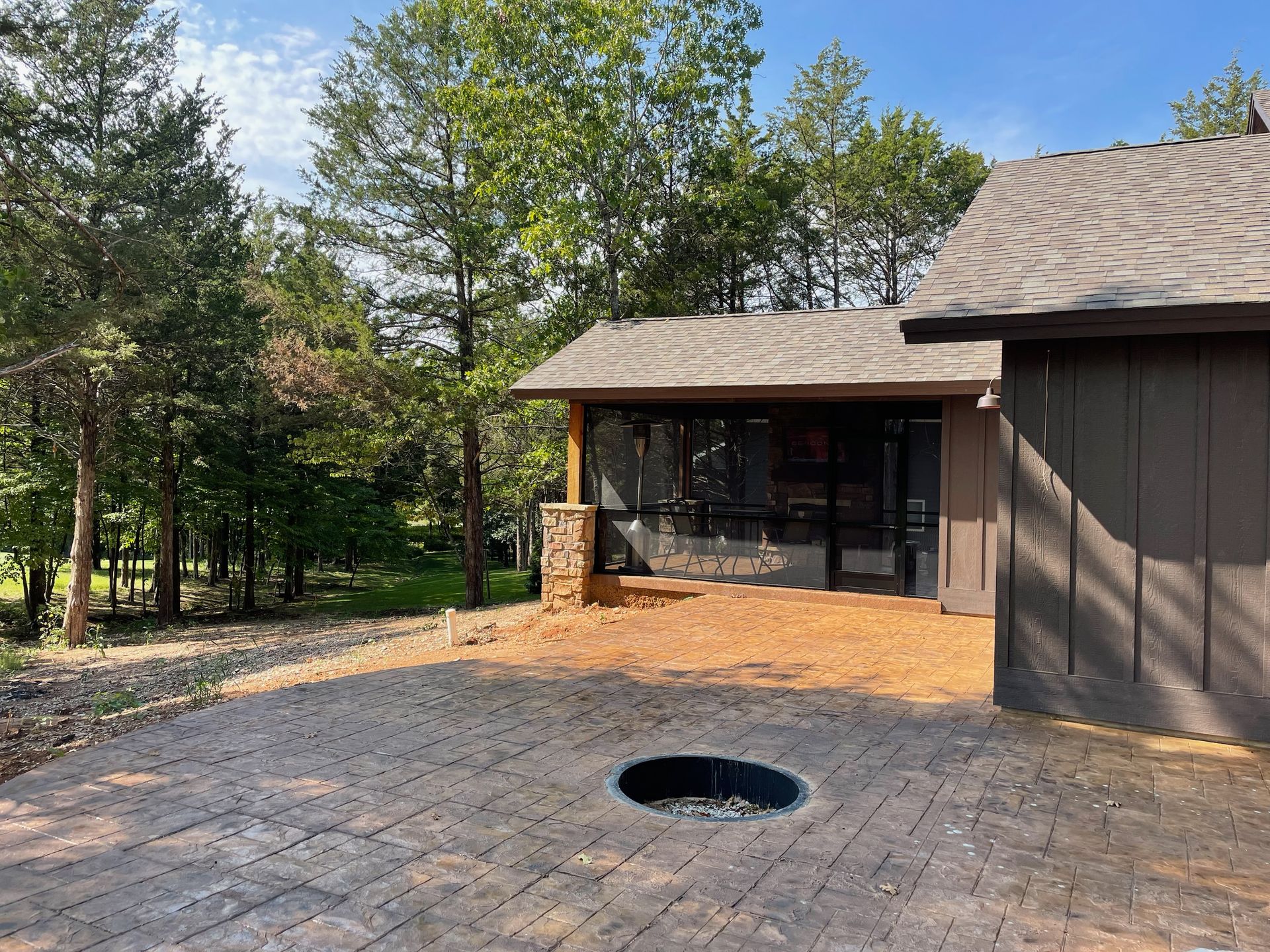 Patio with stone pavers and fire pit beside a brown house, surrounded by trees and greenery