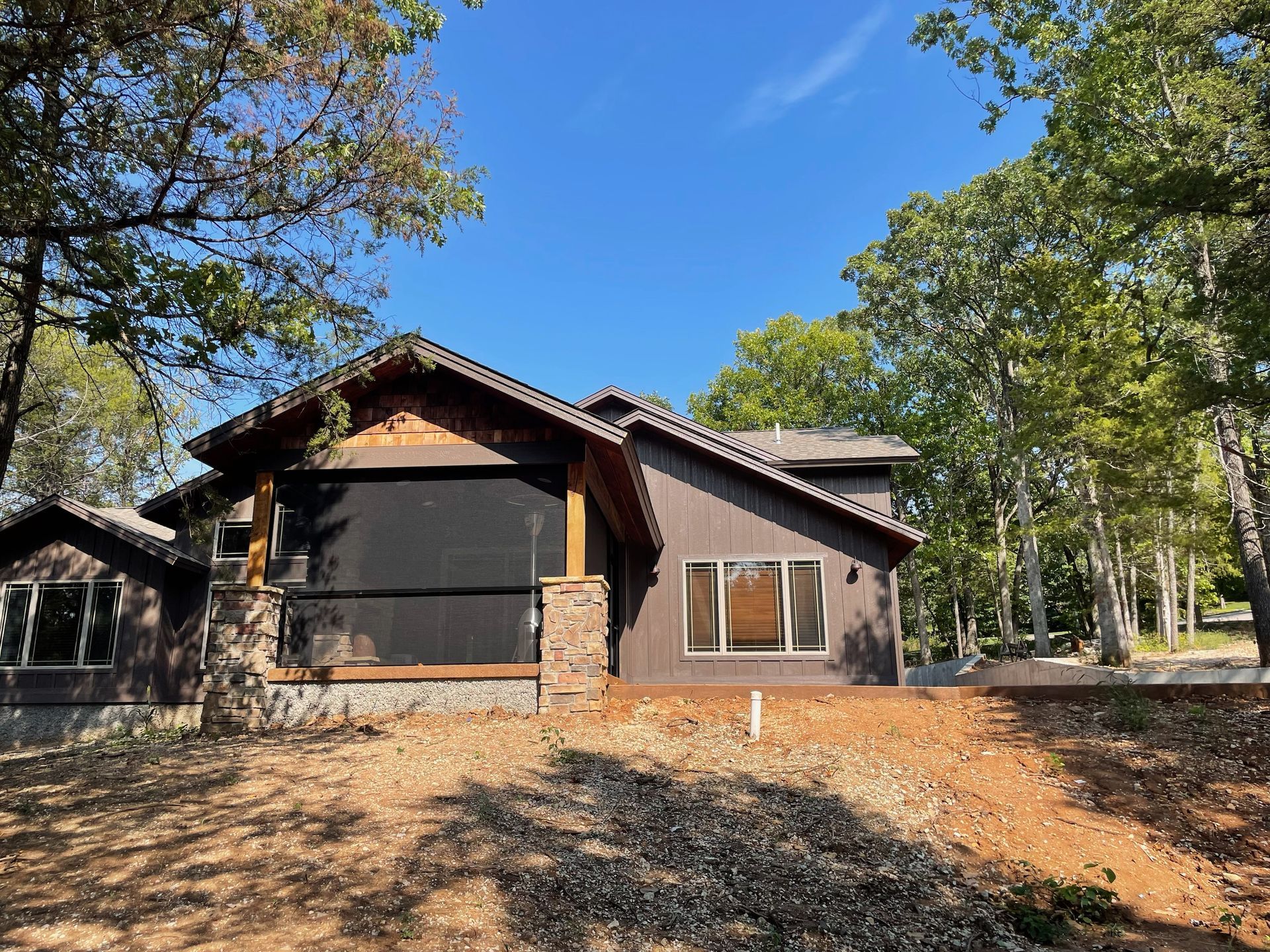 Rustic cabin with dark siding and a stone porch, surrounded by trees under a clear blue sky.
