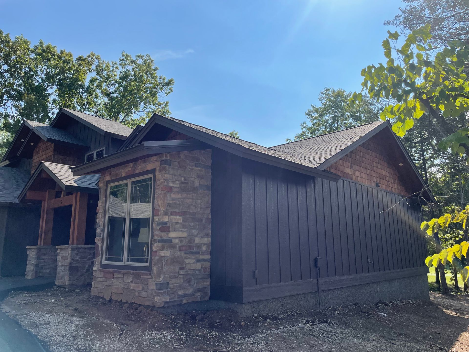 Rustic wooden house with stone exterior and large windows under a bright blue sky, surrounded by trees