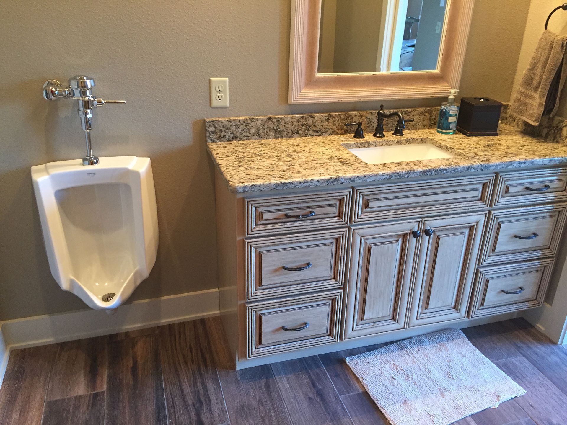 Bathroom with wall-mounted urinal beside a granite vanity with ornate cabinets and a mirror