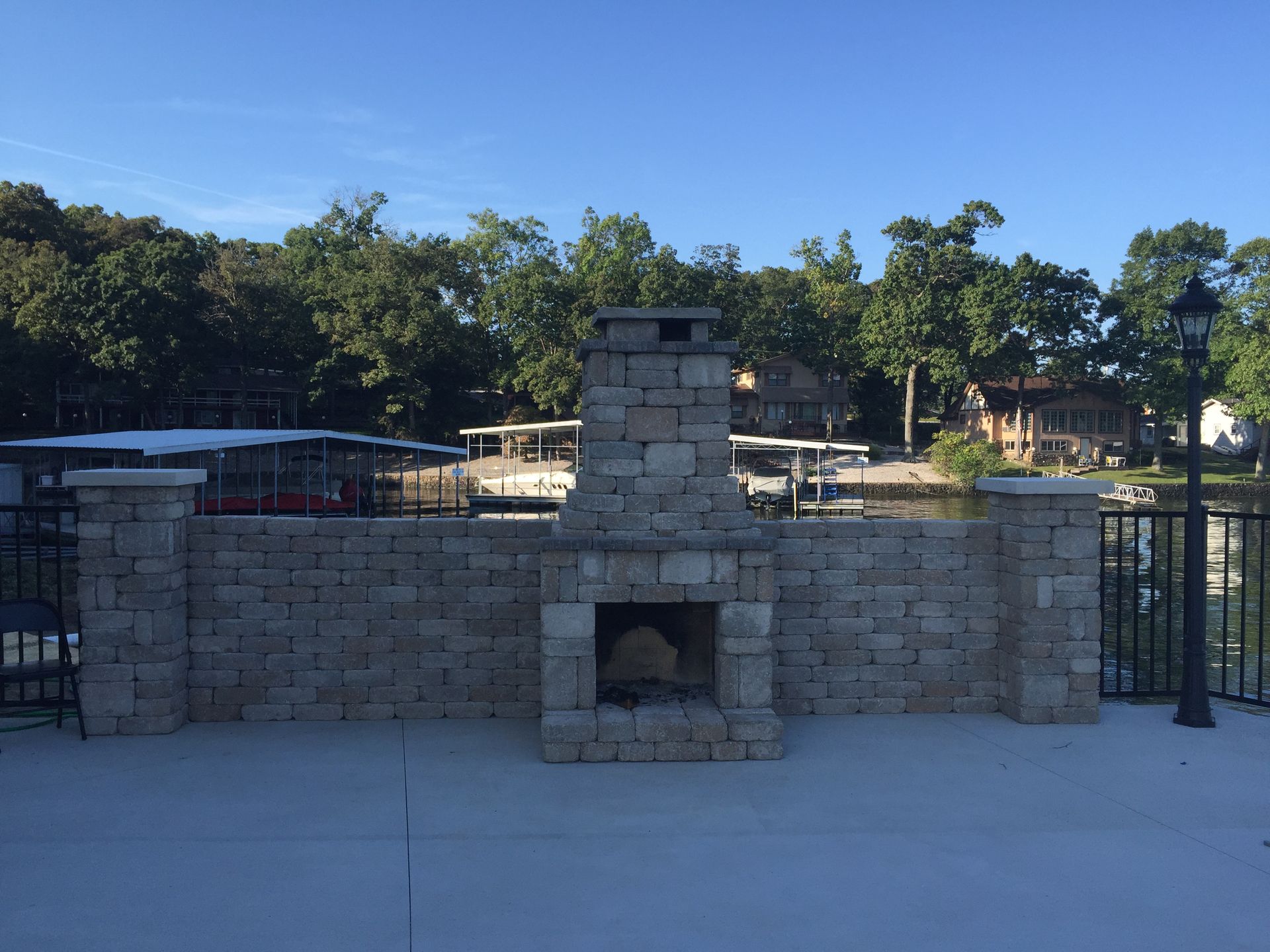 Stone outdoor fireplace and low wall in a sunny park, with trees and a blue sky in the background.