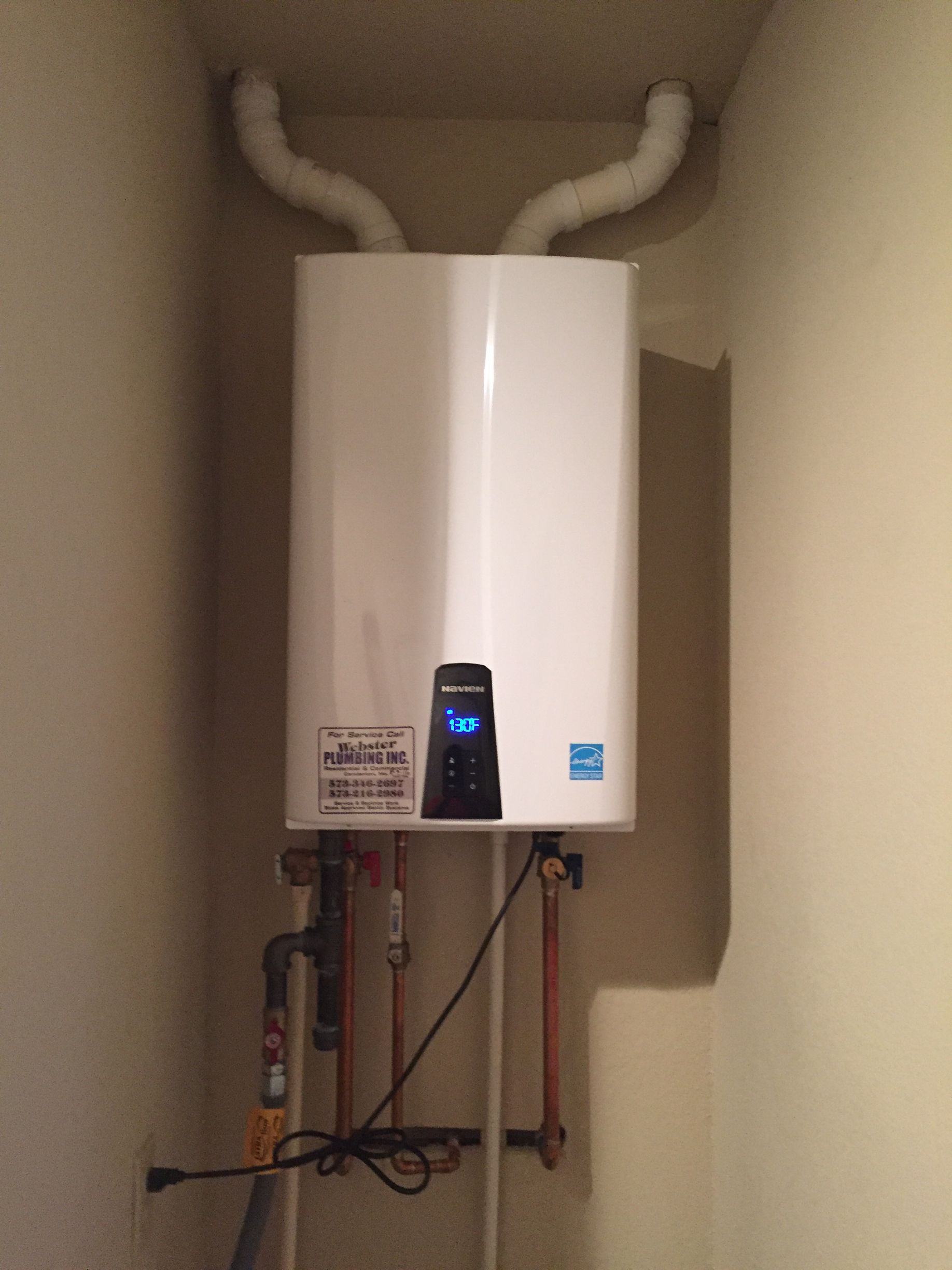 White wall-mounted water heater with exposed pipes and a small digital display in a utility closet