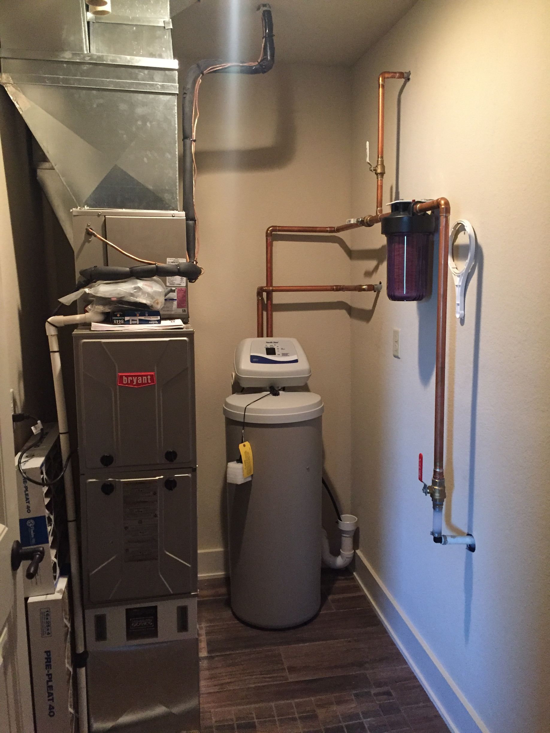 Furnace, water softener, and water heater in a small utility room with exposed ducts and pipes.