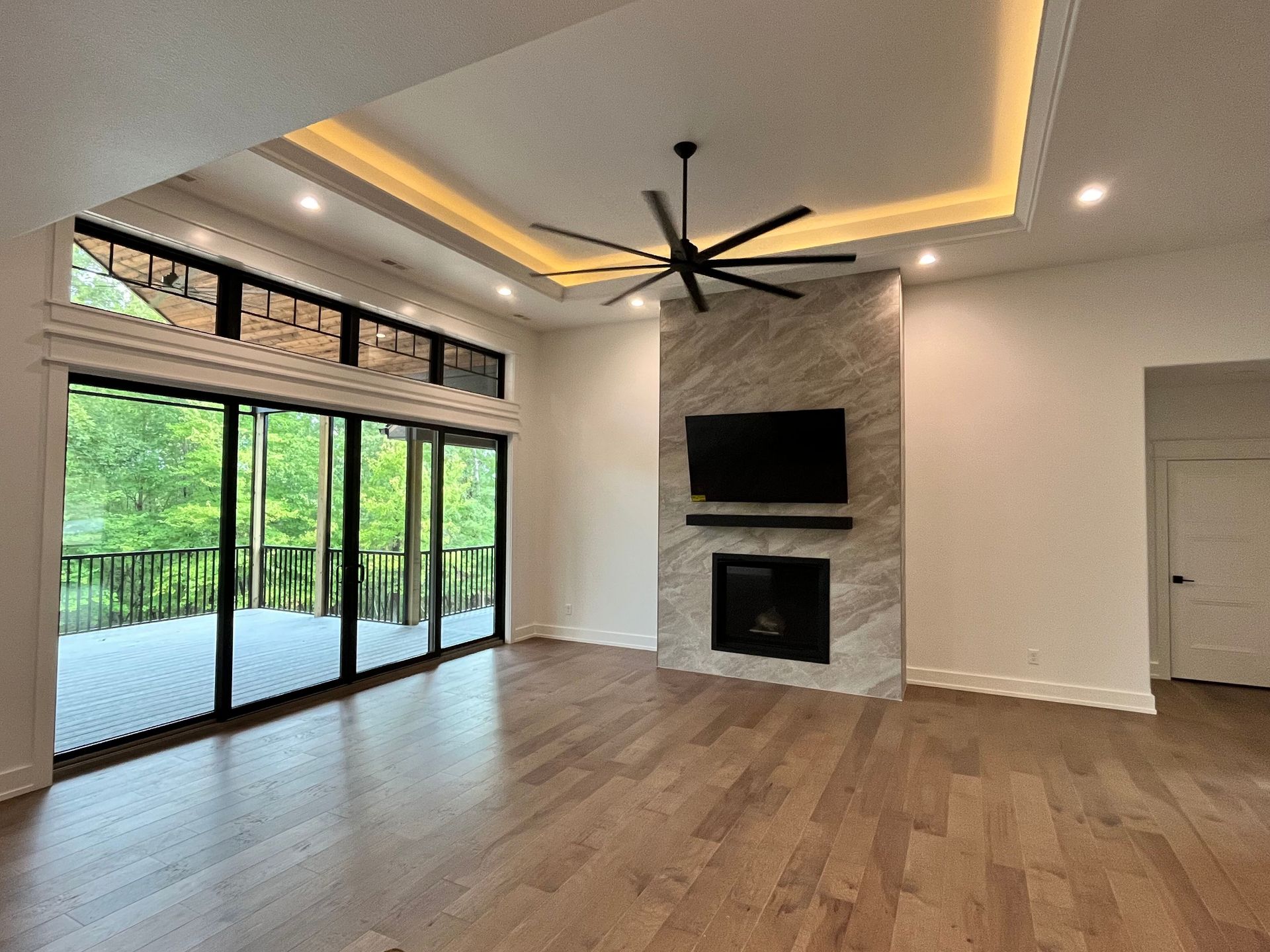 Modern living room with wood floors, fireplace, TV, ceiling fan, and large sliding glass doors to a green deck.