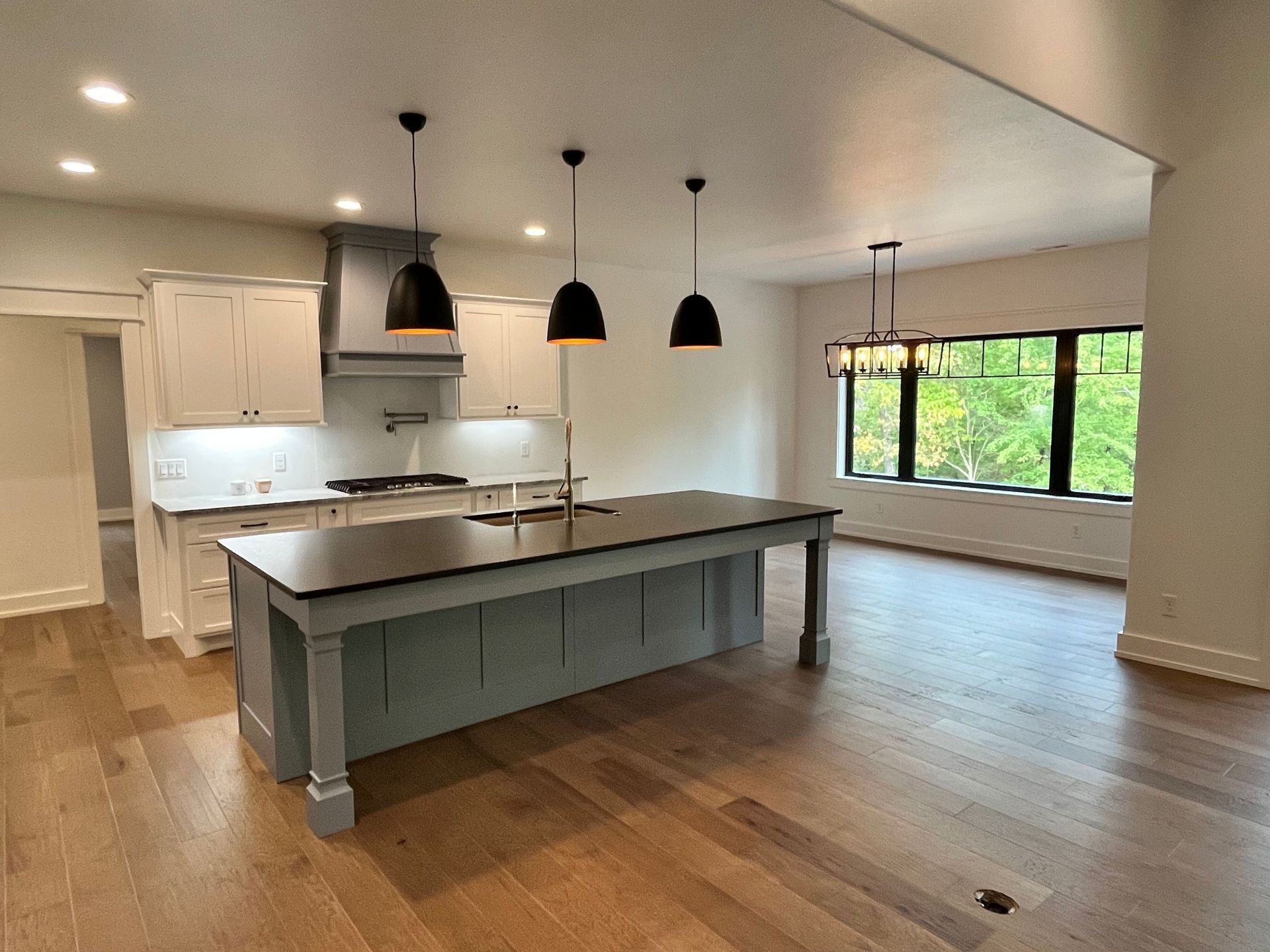Modern kitchen with large gray island, black pendant lights, wood floors, and big windows.