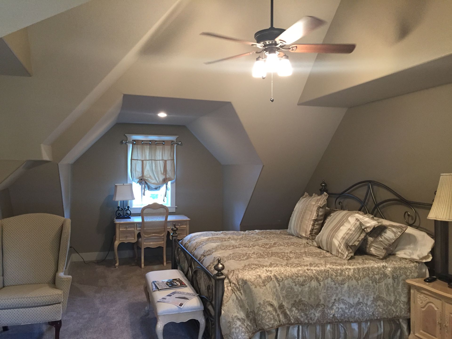 Attic bedroom with bed, chair, desk by window, and ceiling fan-lit sloped ceiling