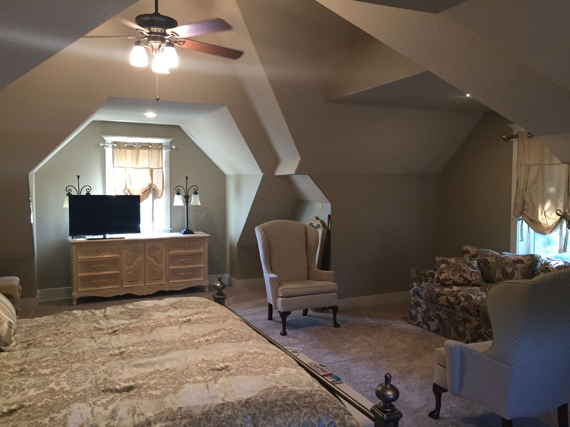Spacious attic bedroom with vaulted ceiling, TV on dresser, and two armchairs on carpeted floors.