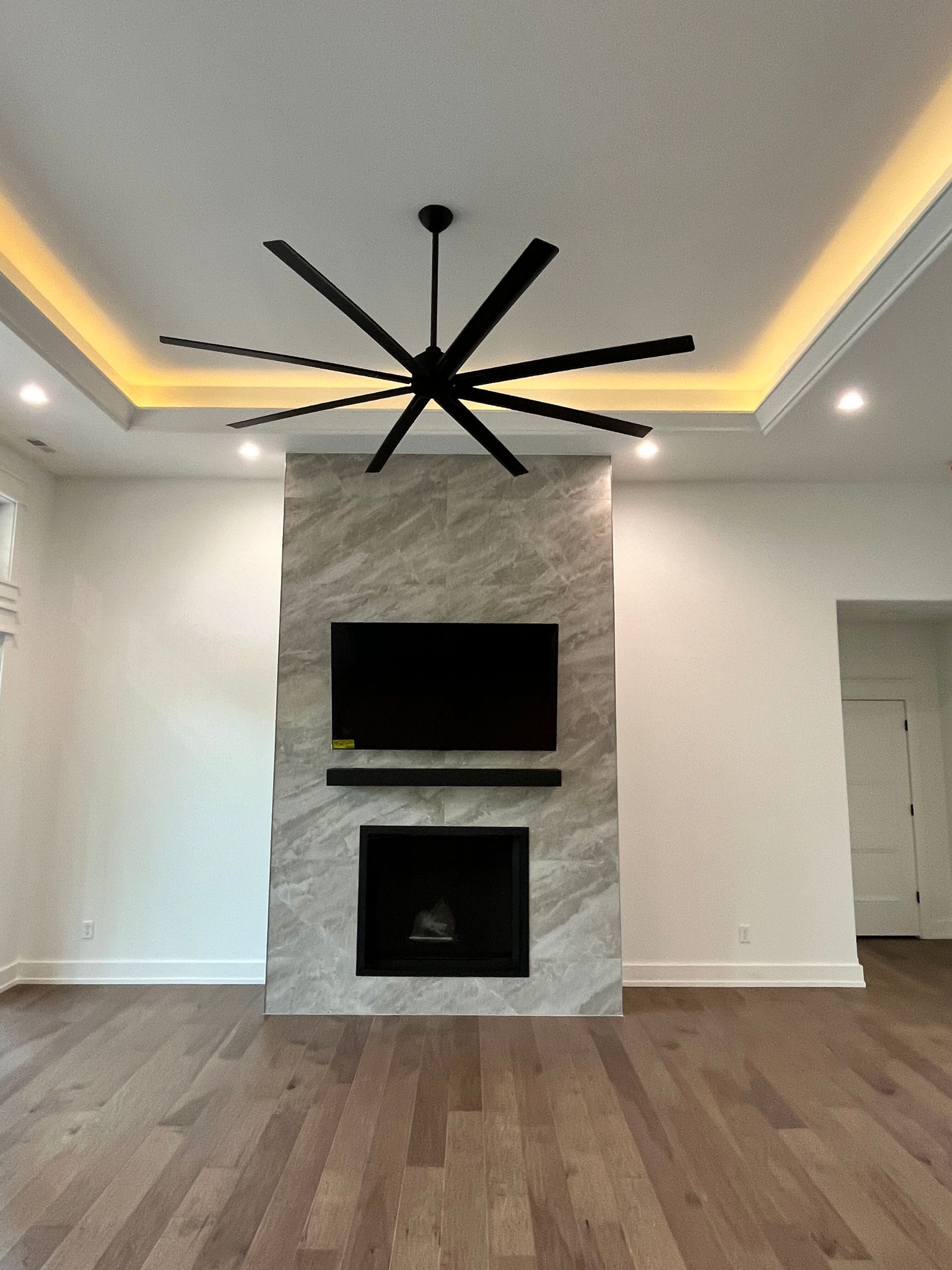 Modern living room with a stone fireplace, recessed ceiling lights, wood floors, and a large black ceiling fan.