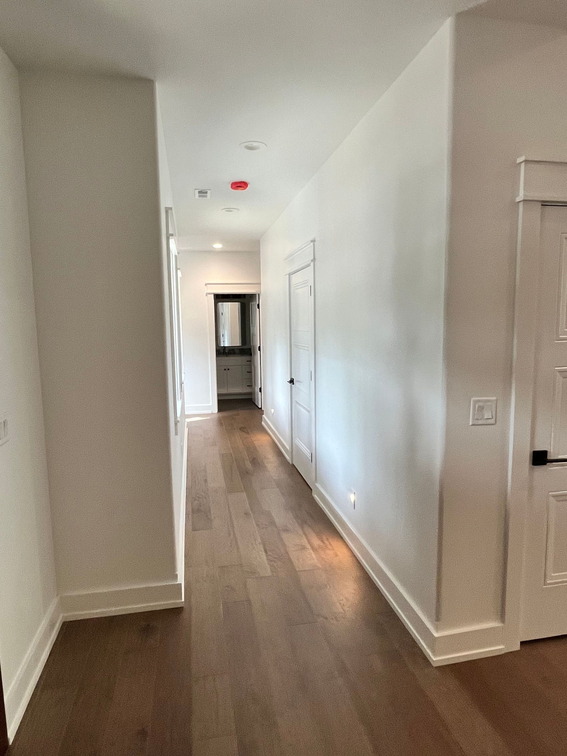 Bright white hallway with wood floors and several doors, leading to a window at the far end.