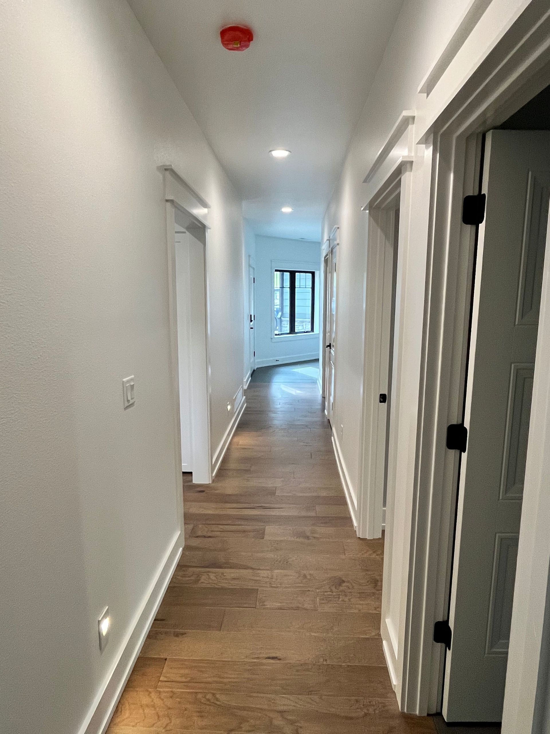Narrow white hallway with wood floors, doors on both sides, and a window at the far end.