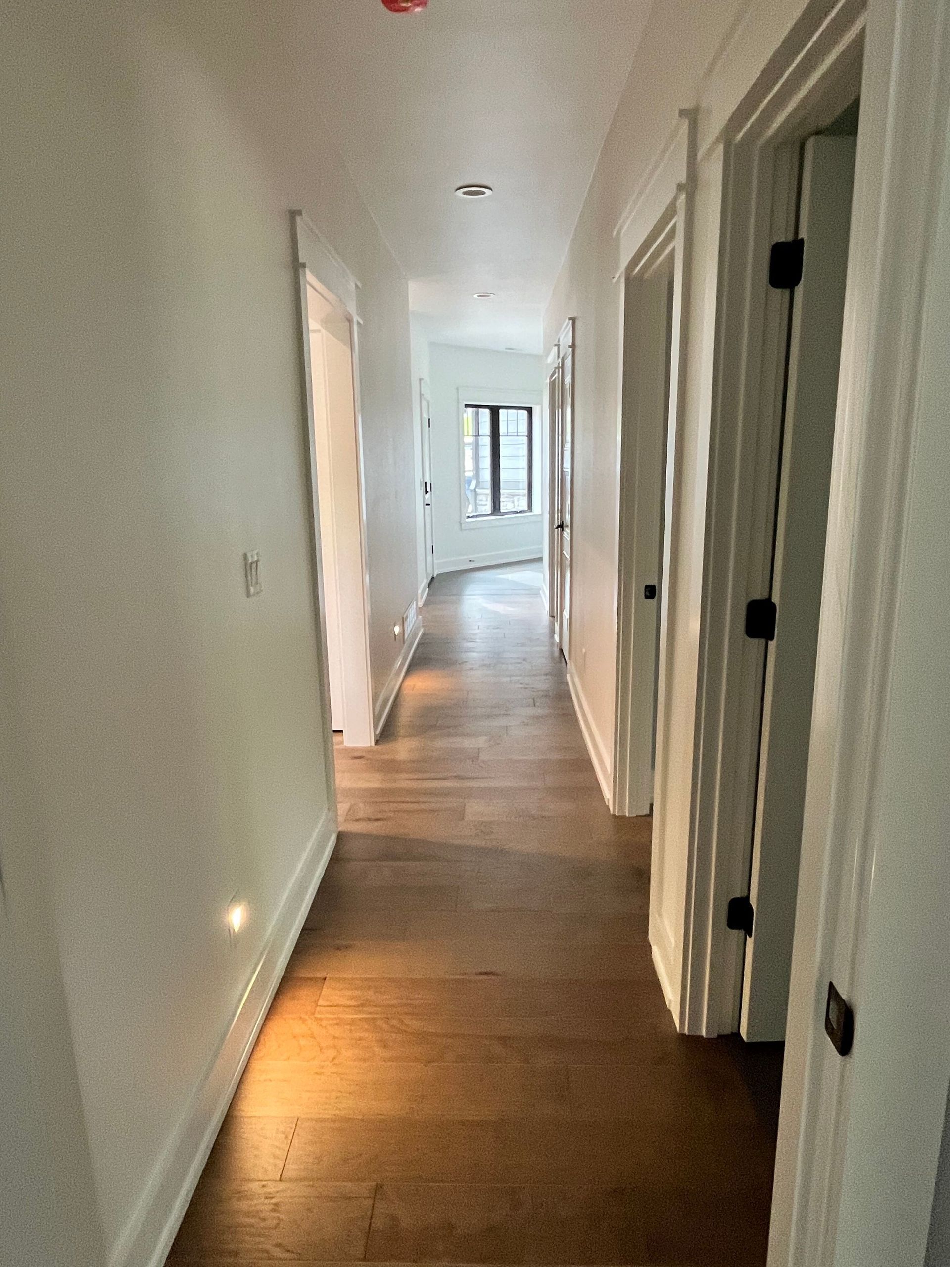 Narrow white hallway with wood floors and several open doorways, lit by daylight from the far end.