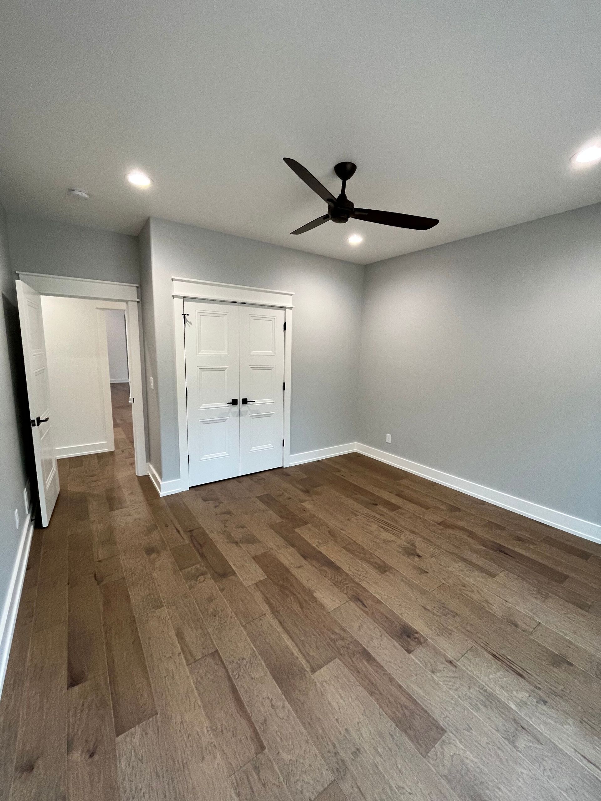 Empty gray bedroom with wood floors, white closet doors, ceiling fan, and doorway to hall
