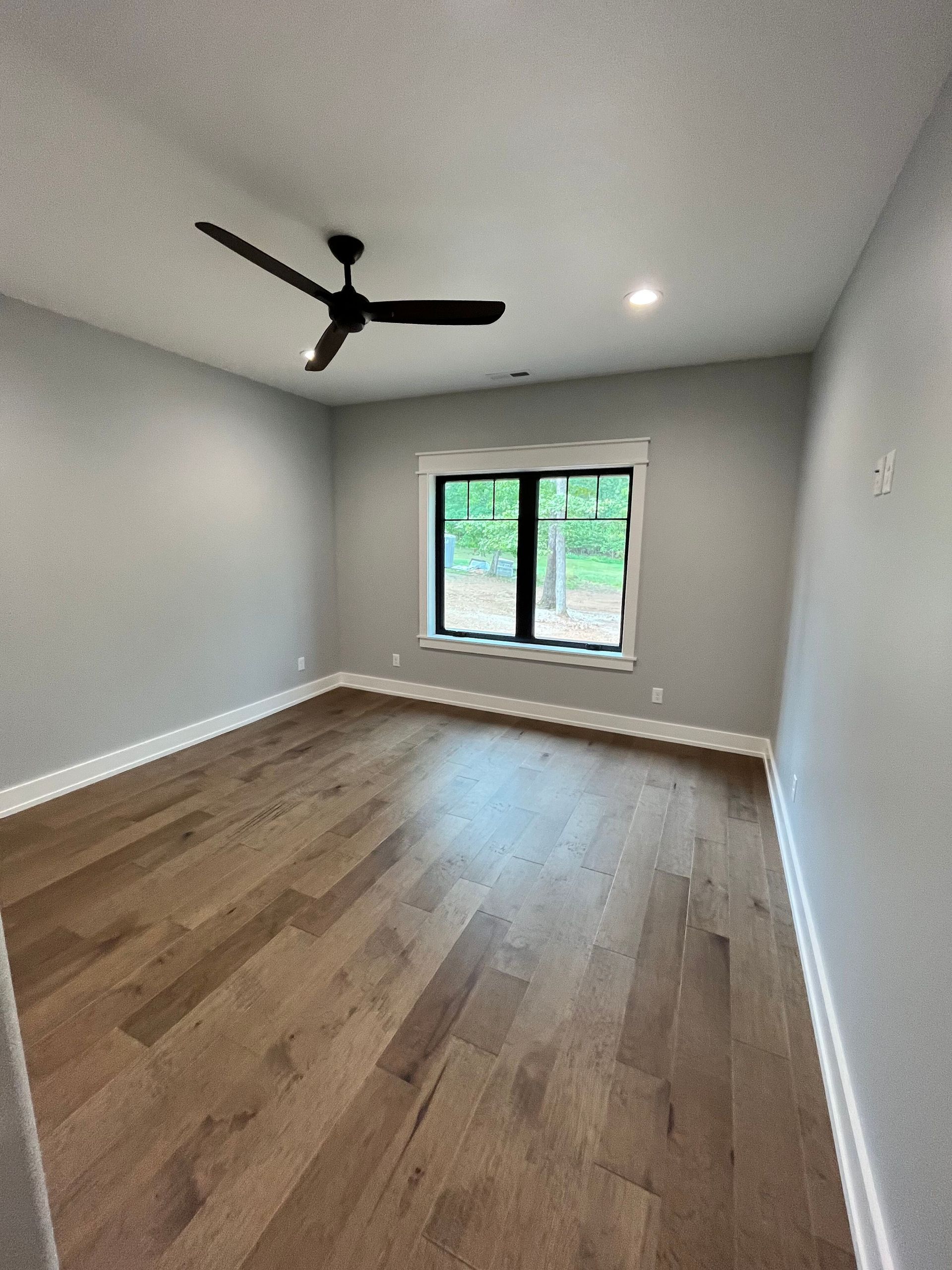 Empty gray bedroom with wood floors, ceiling fan, and window overlooking greenery.