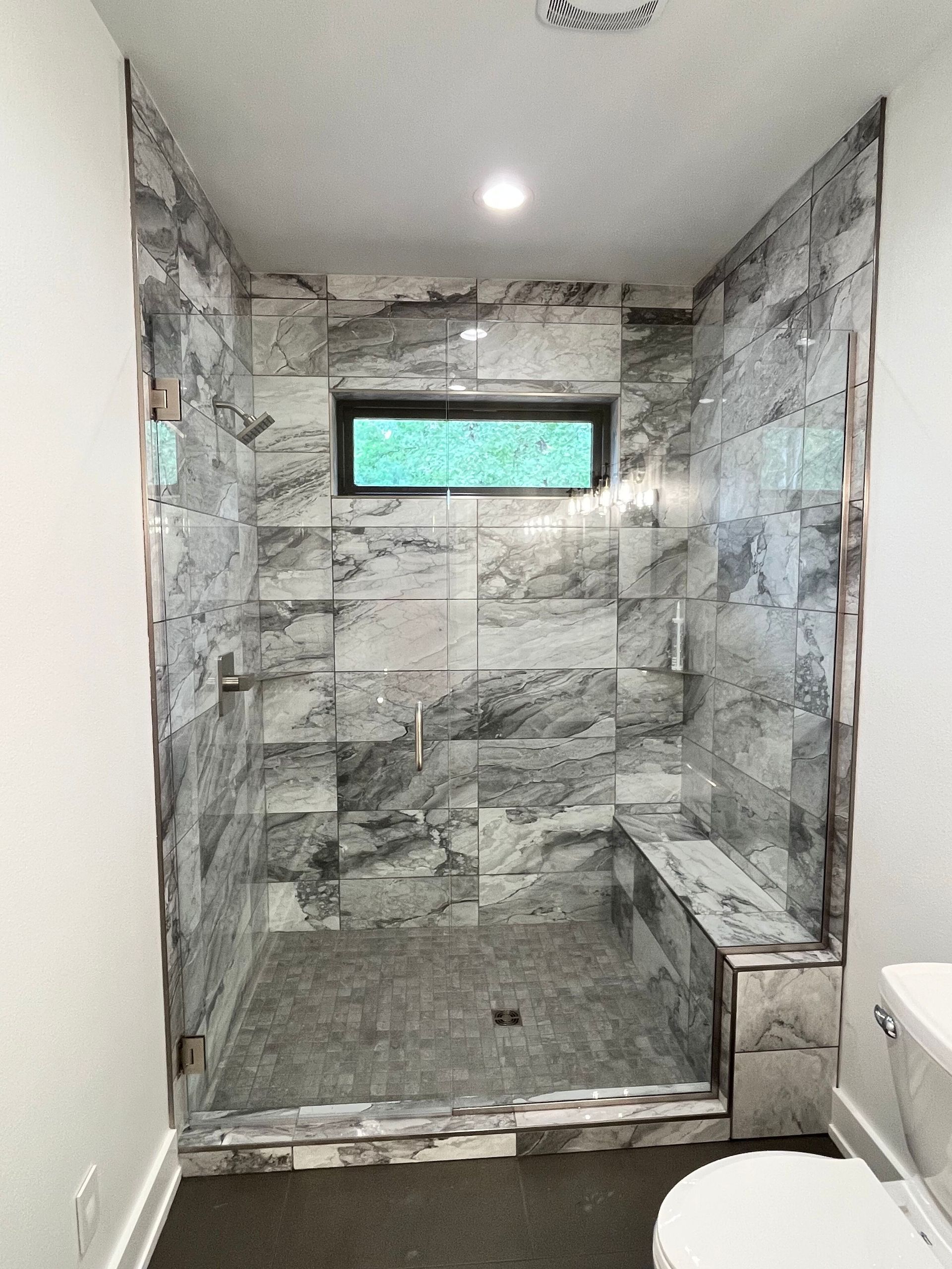 Modern tiled walk-in shower with a small frosted window and adjacent toilet in a bathroom