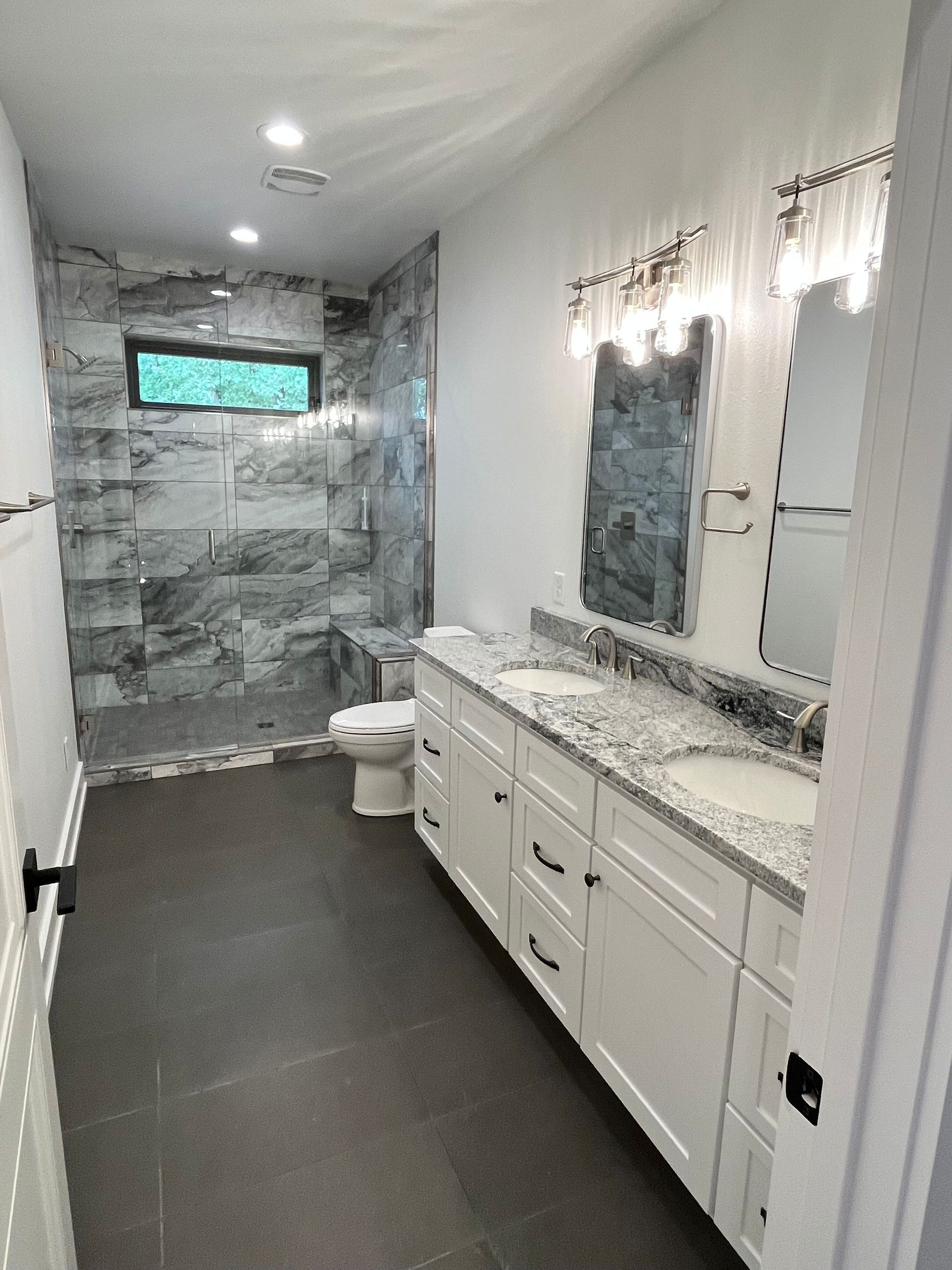 Long, narrow bathroom with white vanity, marble tile, mirrors, and a stone-tiled shower at the far end.