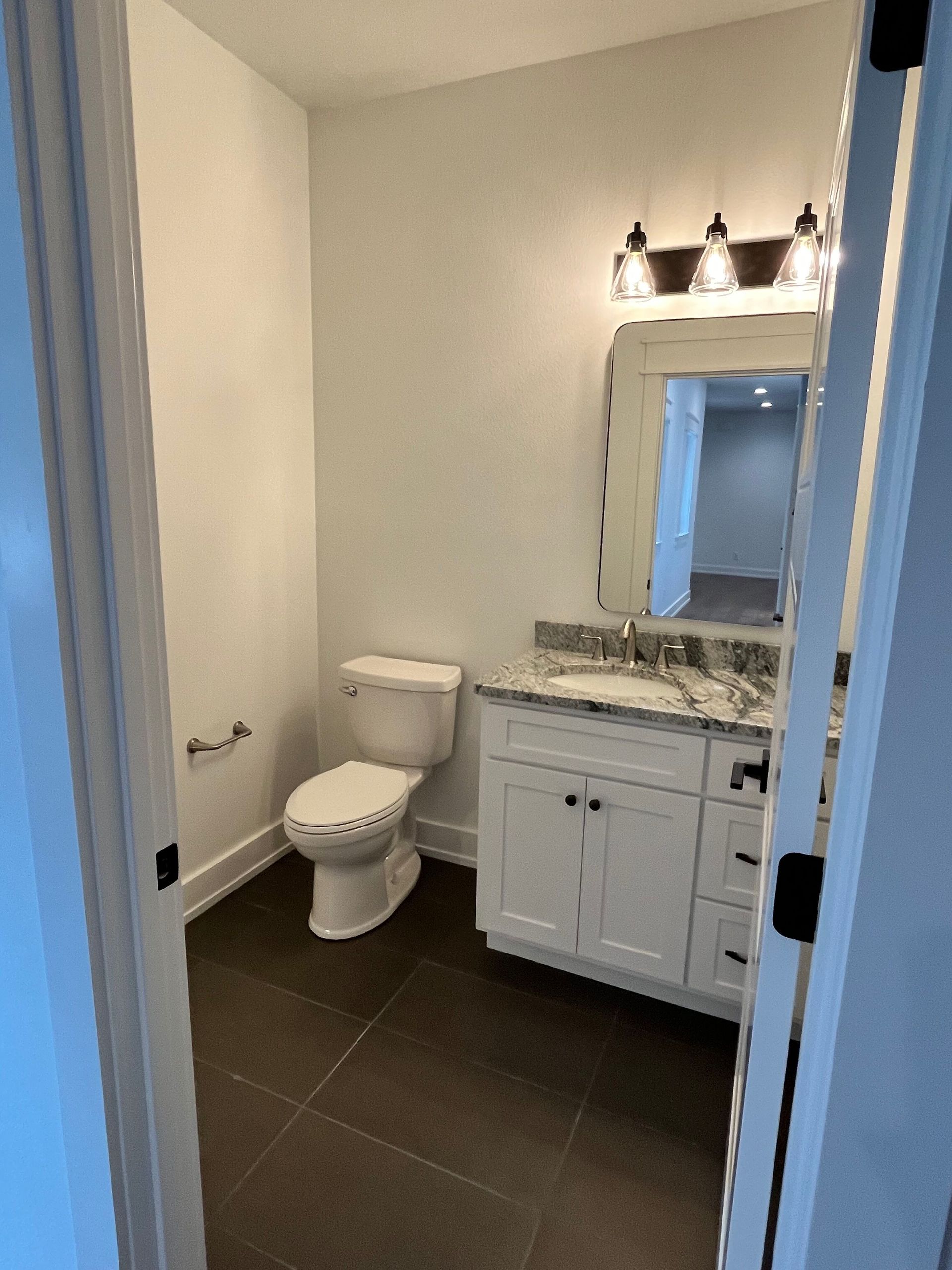Small bathroom with white vanity, toilet, mirror, and dark tile floor.
