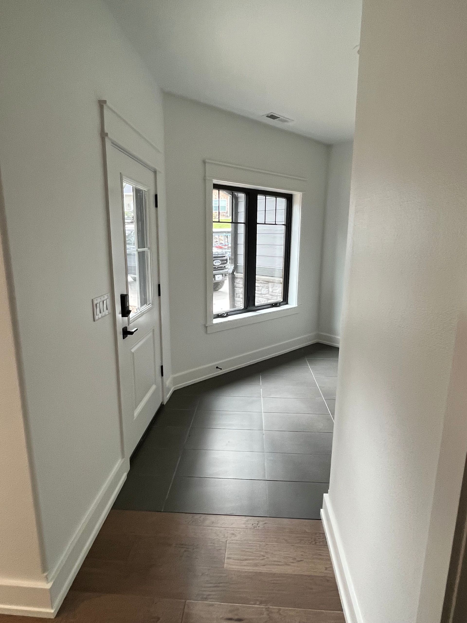 Narrow hallway with white walls, wood and dark tile flooring, and a window at the end.