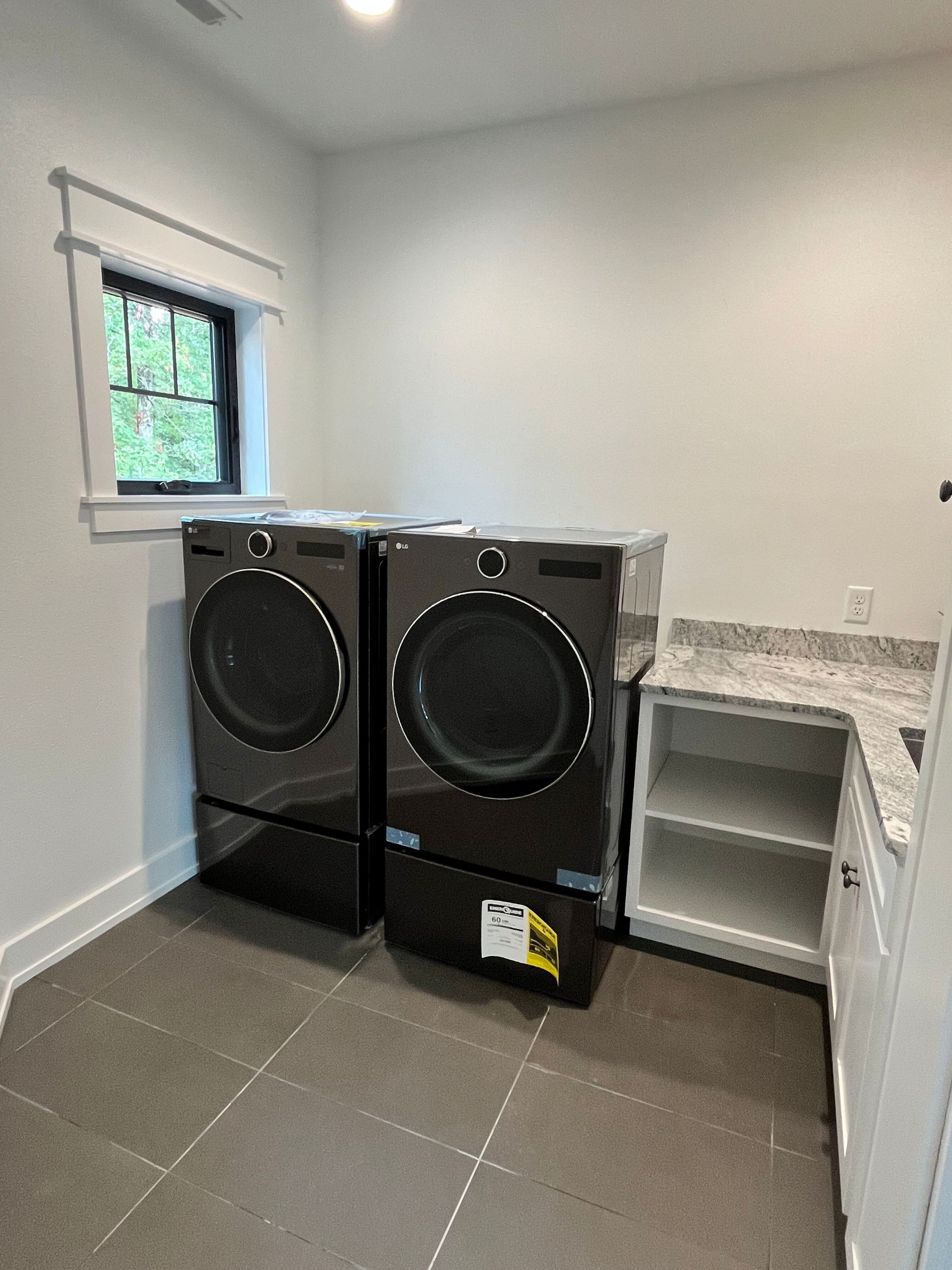 Laundry room with two black front-load washers beside a small window and white shelves.