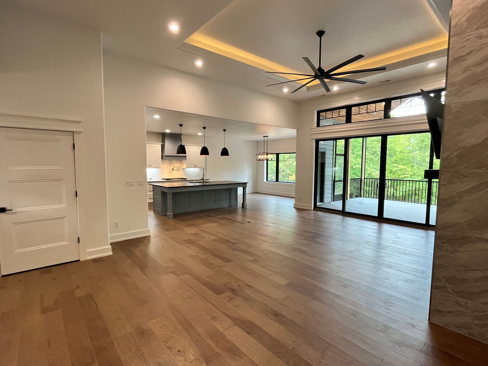 Empty modern open-plan living room and kitchen with wood floors, white walls, large glass doors, and ceiling fan
