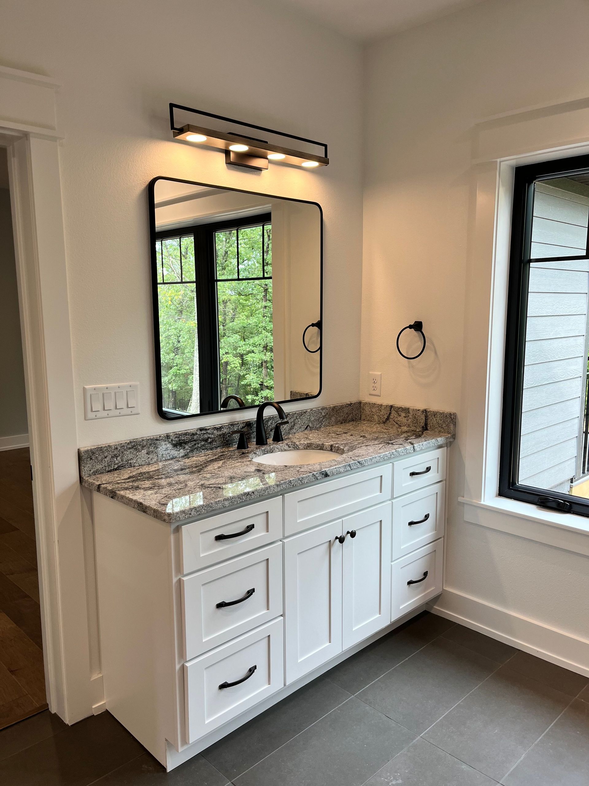 White bathroom vanity with granite countertop, mirror, and black-framed windows in a bright room