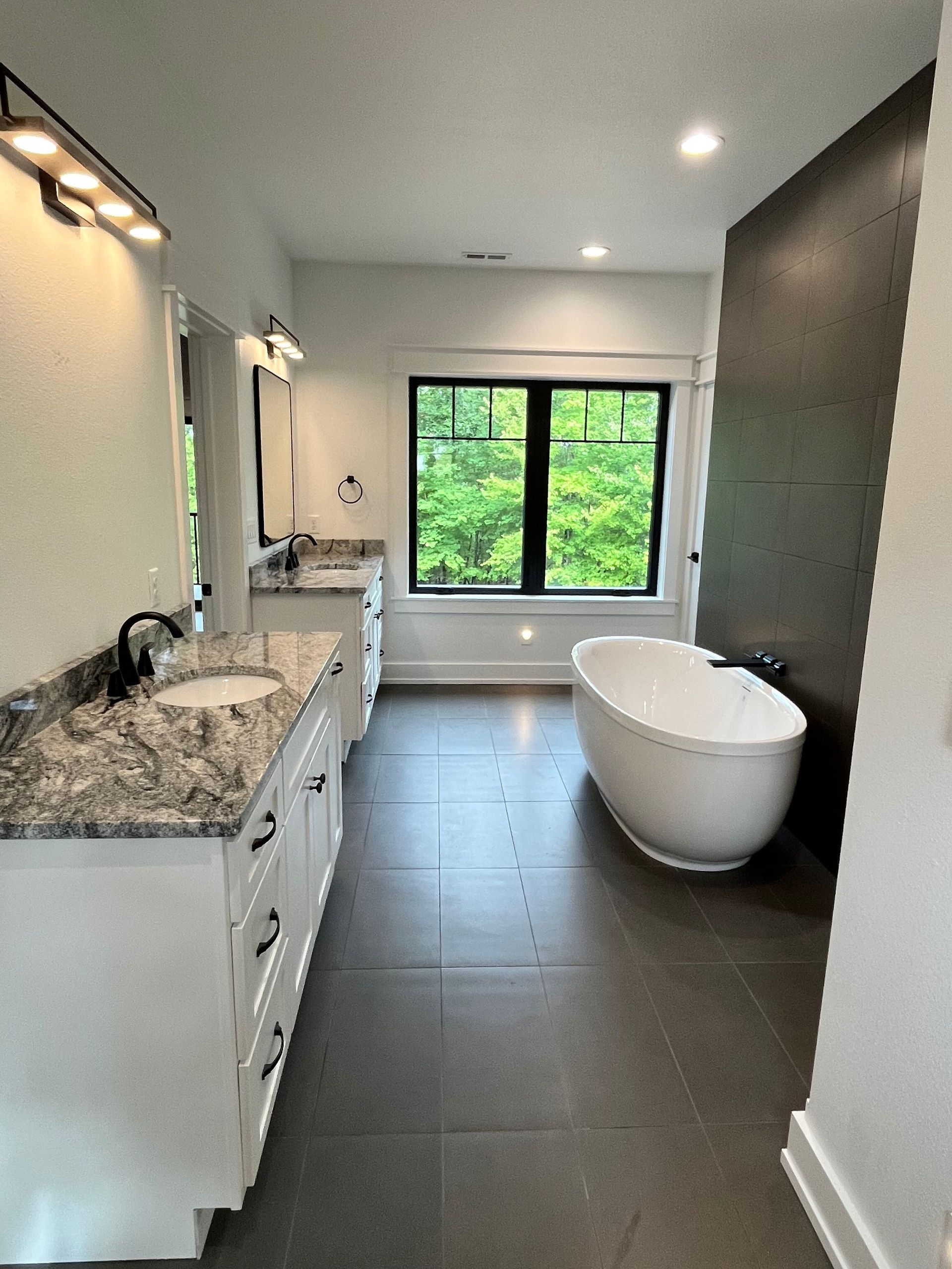 Modern bathroom with floating vanity, freestanding tub, dark tile floor, and large window overlooking greenery