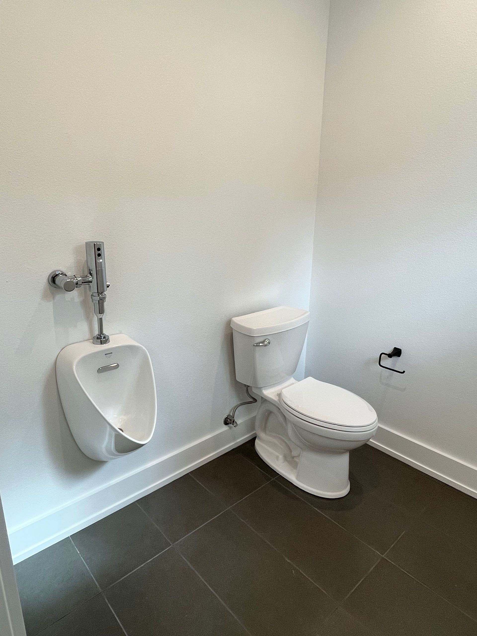 Small tiled restroom with a urinal and toilet against white walls and dark floor tiles