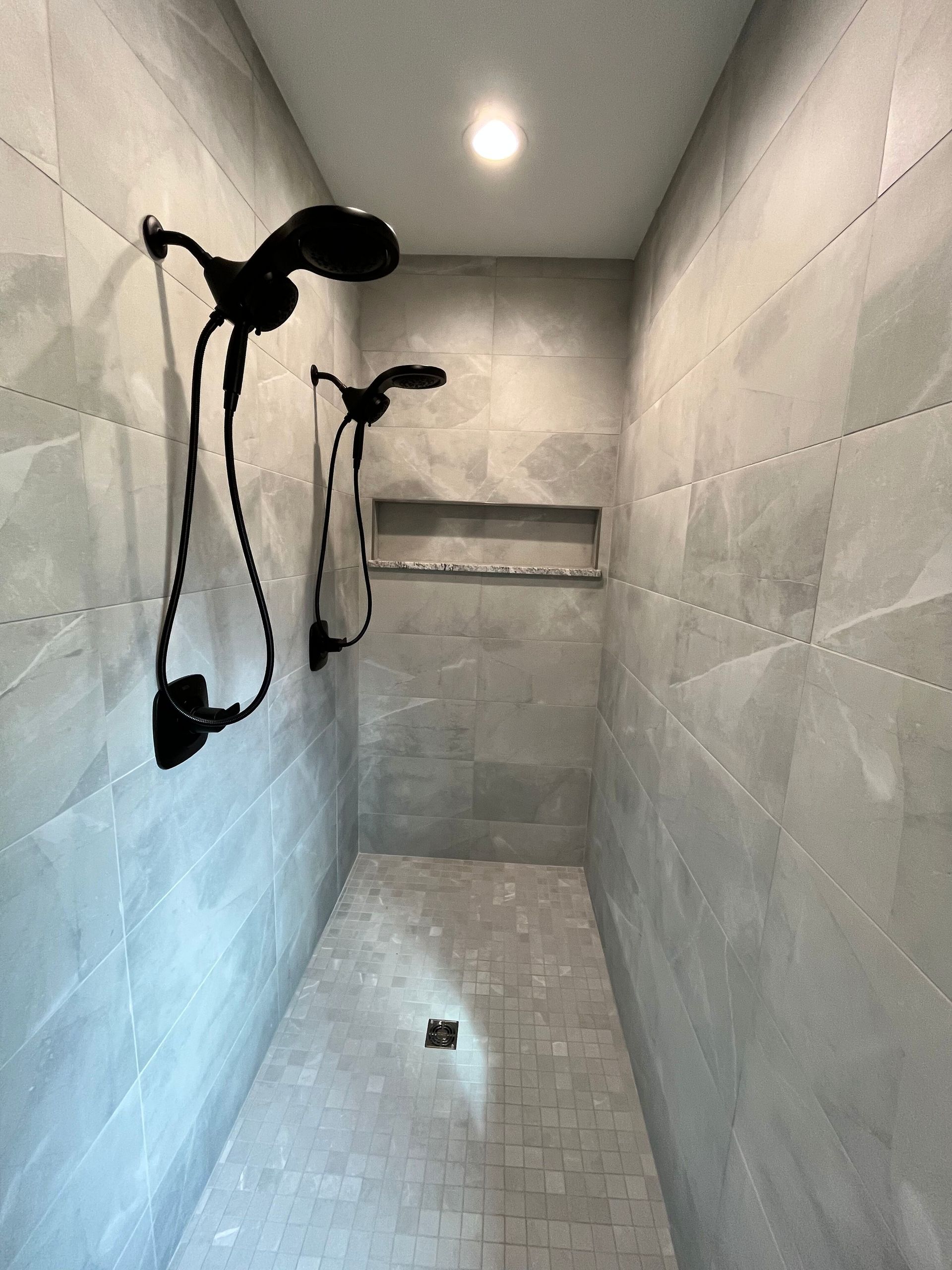 Tiled shower stall with two black shower fixtures, a shelf, and a ceiling light.