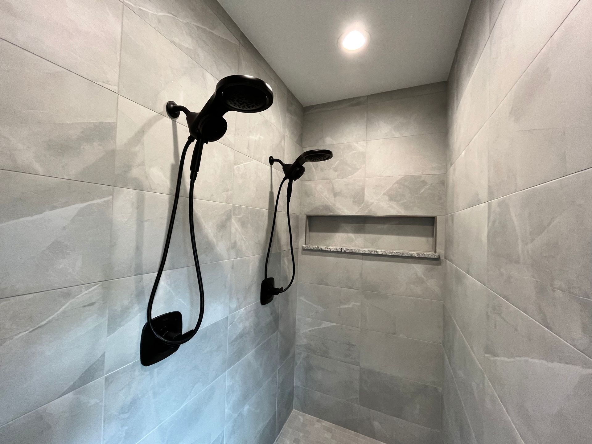 Gray tiled shower with two black showerheads and a built-in shelf.