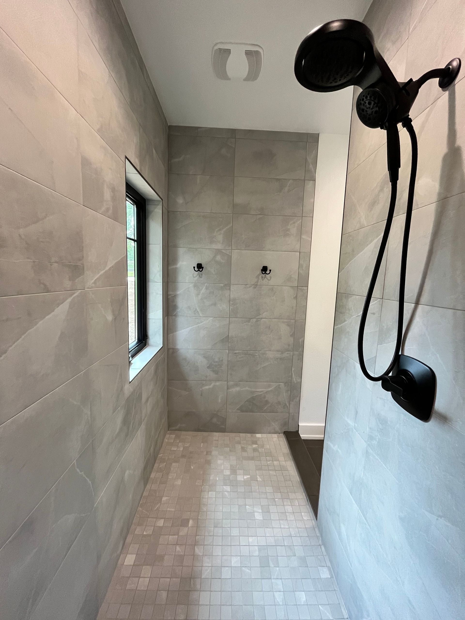 Modern tiled shower with black handheld showerhead and window on the left.