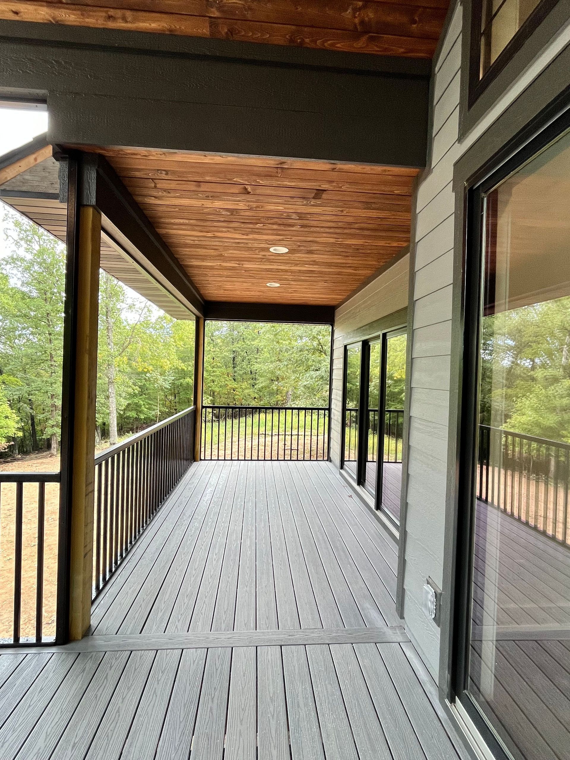 Covered wooden porch with railing, slatted floor, and large windows overlooking trees