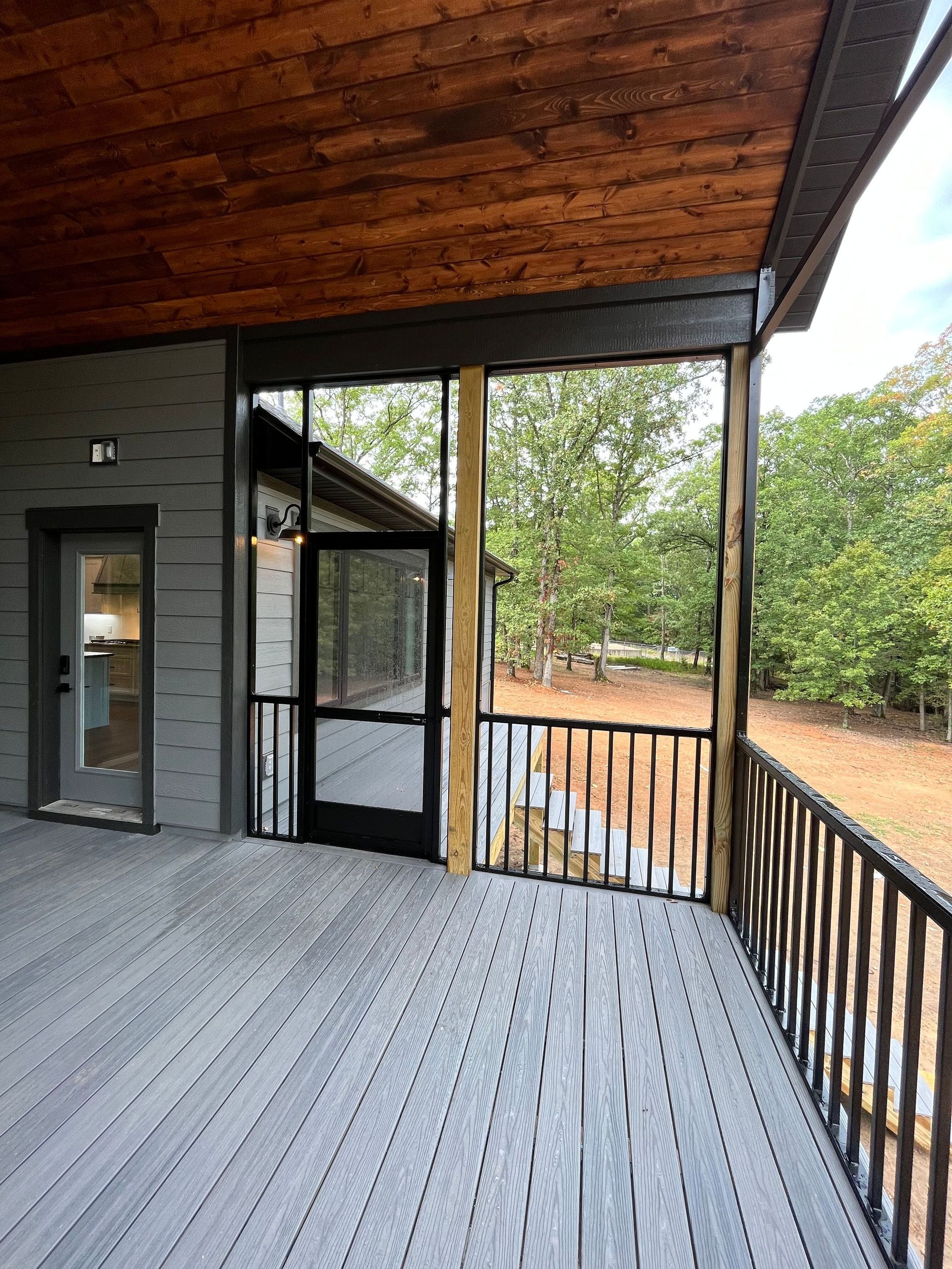 Covered wooden porch with railing, gray deck, and trees overlooking a dirt yard