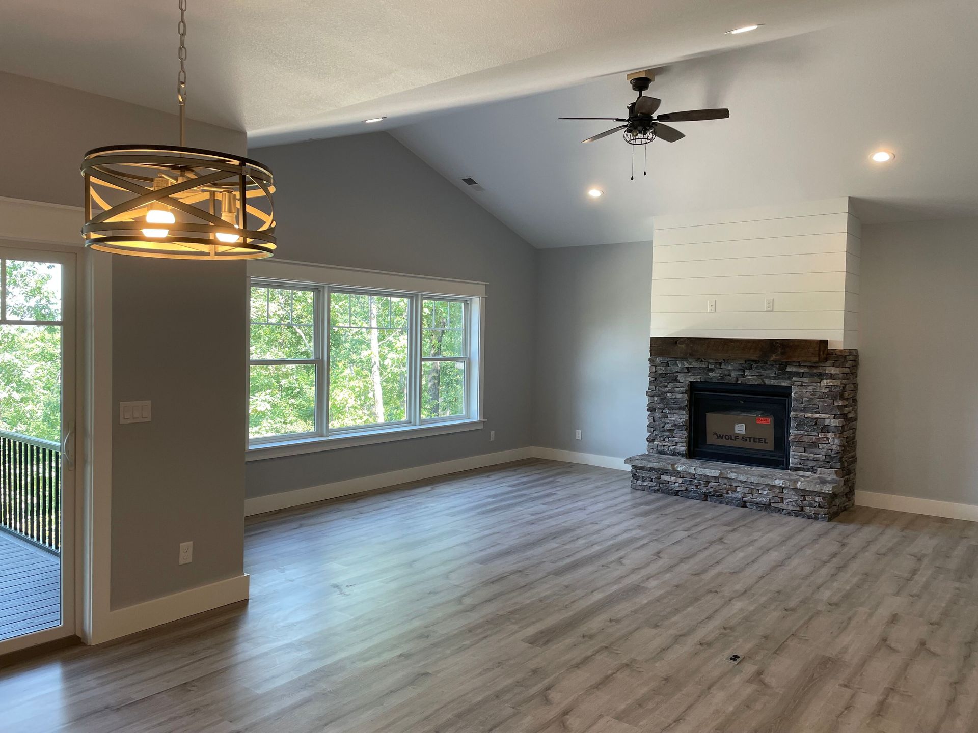 Empty living room with gray walls, wood floors, large windows, and a stone fireplace.