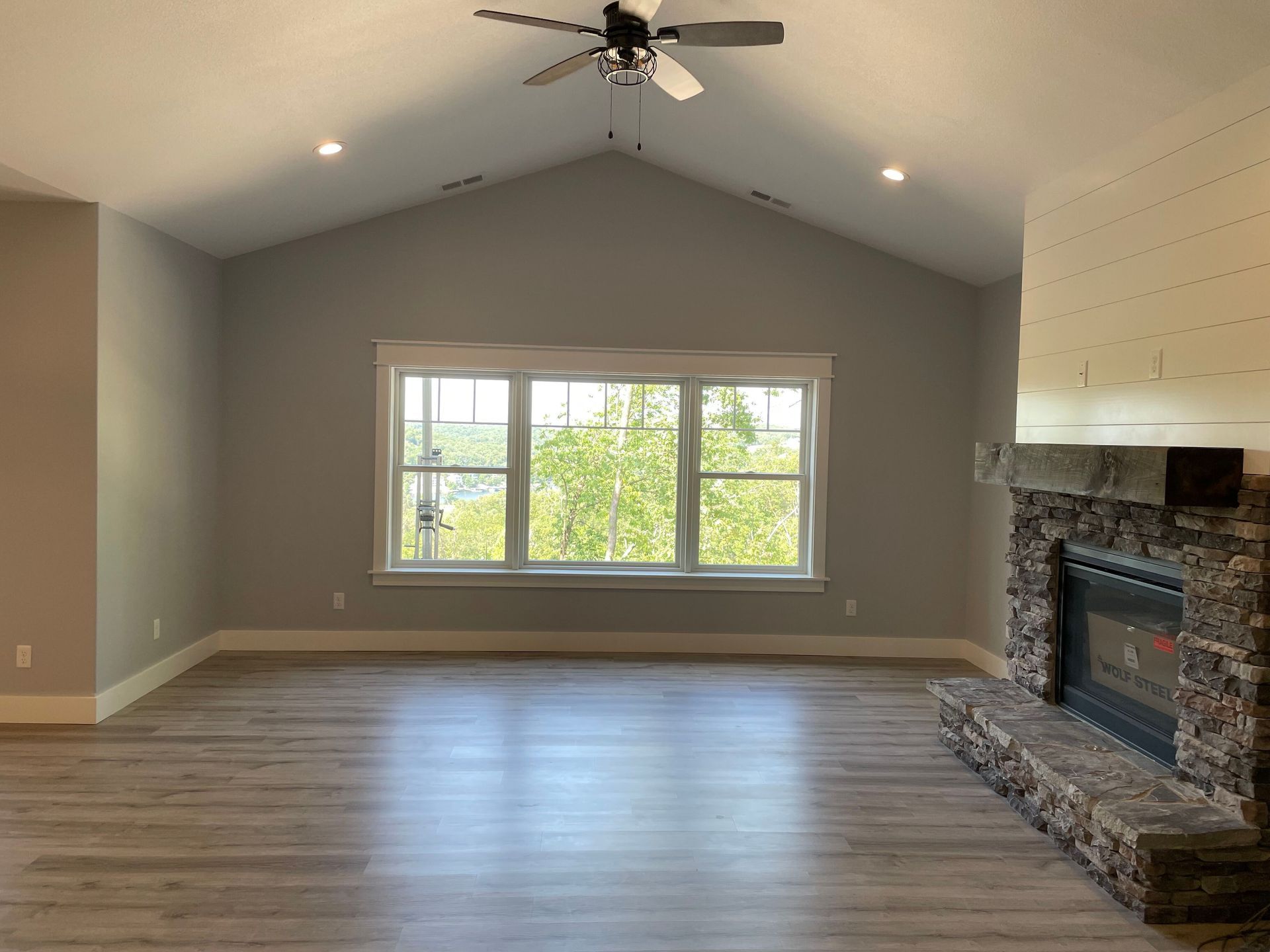 Empty living room with vaulted ceiling, large window, and stone fireplace