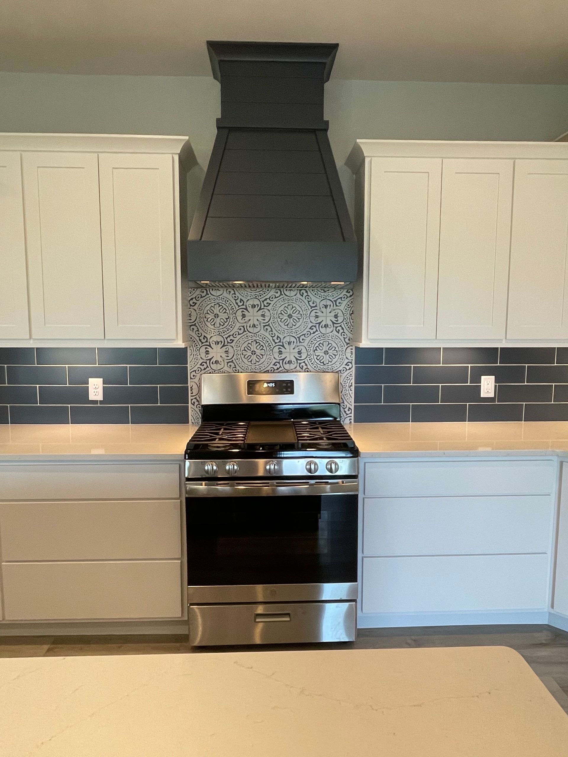 Modern kitchen with white cabinets, black range hood, stainless stove, and blue lower cabinets against tiled backsplash