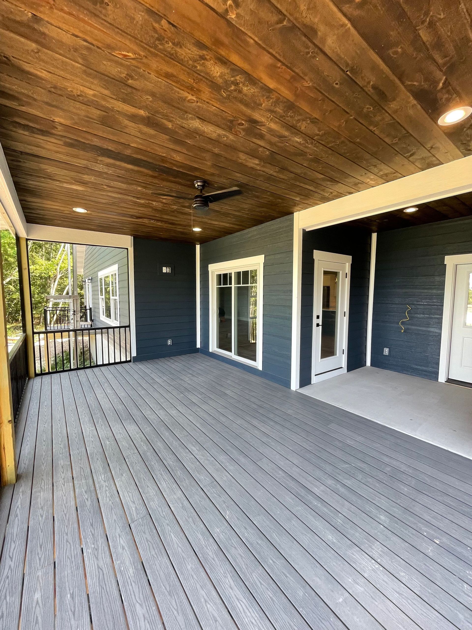Covered porch with gray decking, wood plank ceiling, black siding, and white-trimmed doors and windows.