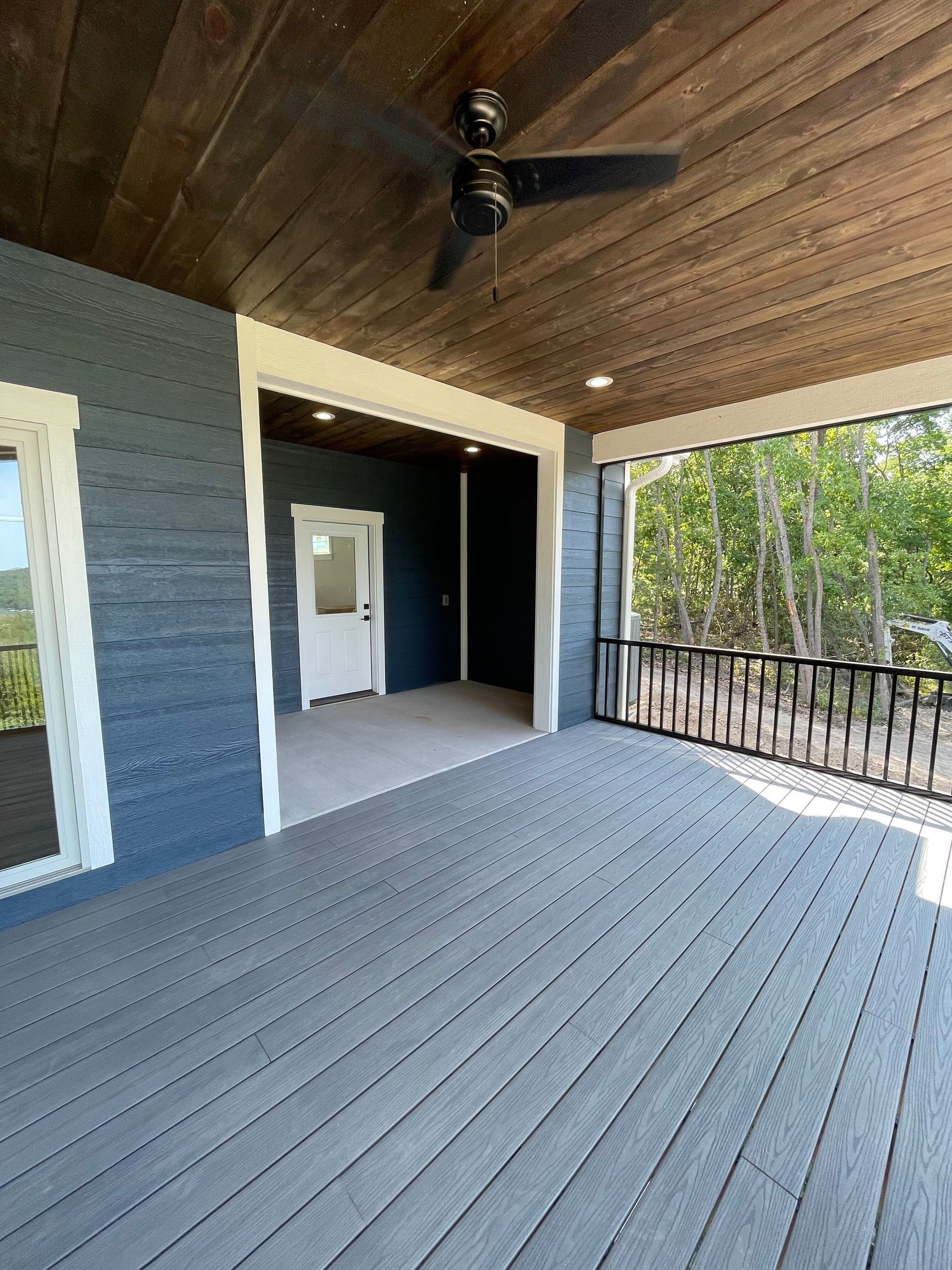 Covered wooden porch with gray deck, dark ceiling fan, white-trimmed doorway, and railing overlooking trees