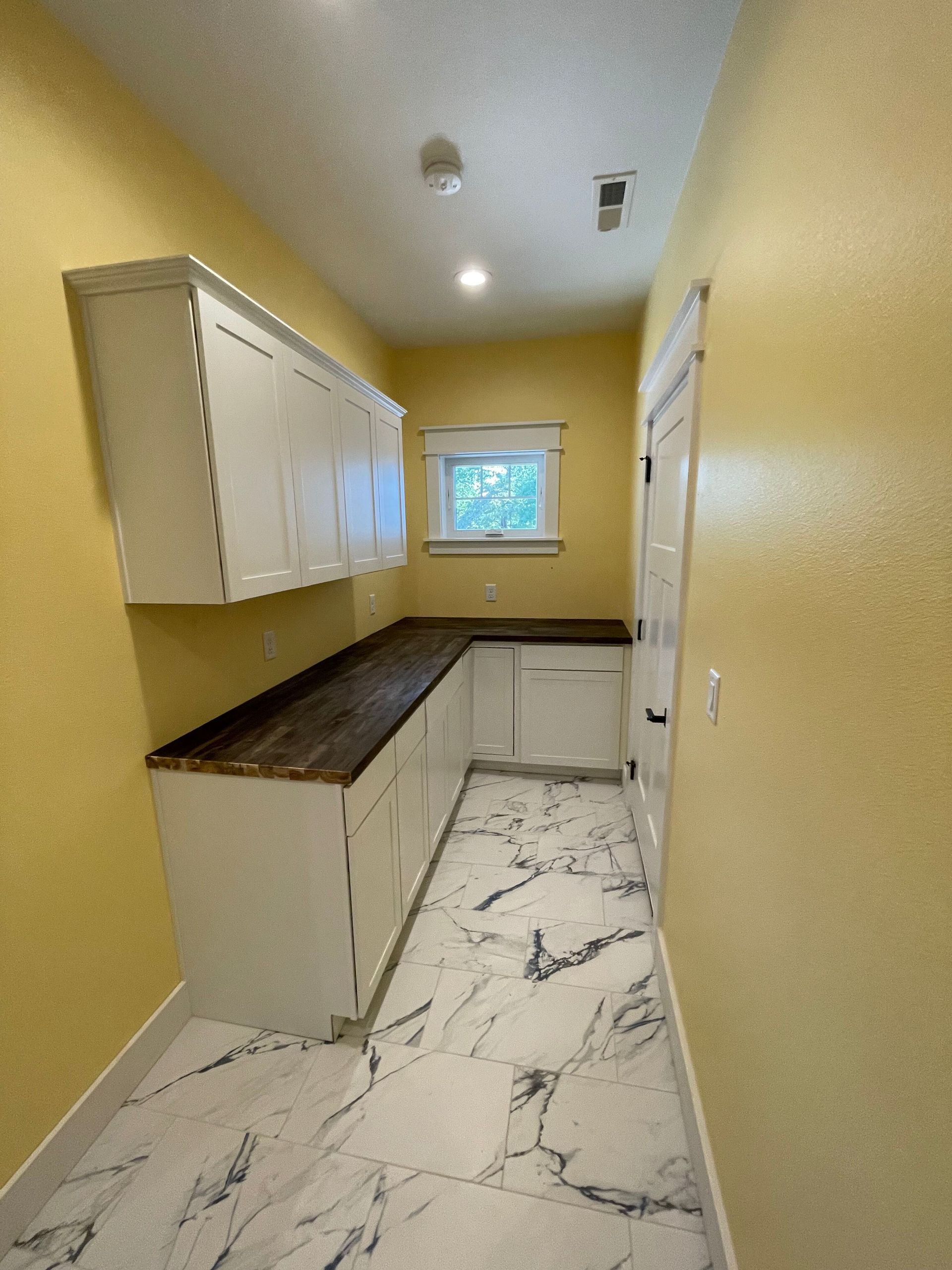 Narrow yellow kitchen with white cabinets, dark counters, marble floor, and a small window at the end.