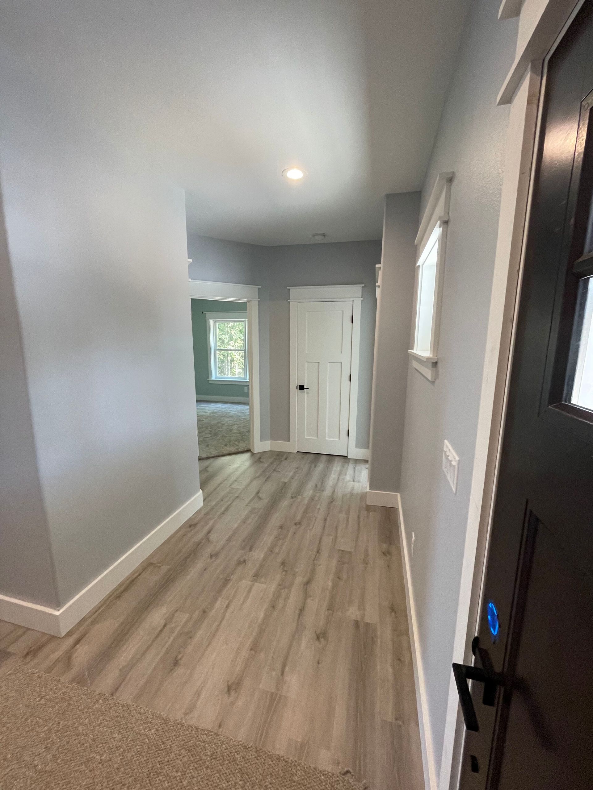 Empty hallway with gray walls, wood-look flooring, and white doors leading to a bright room.
