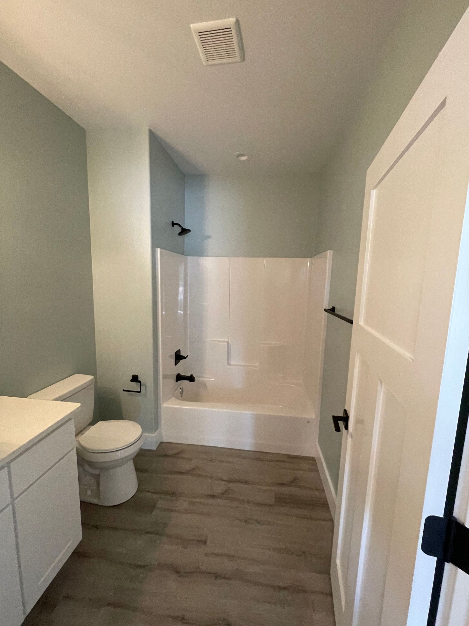 Small bathroom with white tub, toilet, sink, and light wood floor, viewed from the doorway.